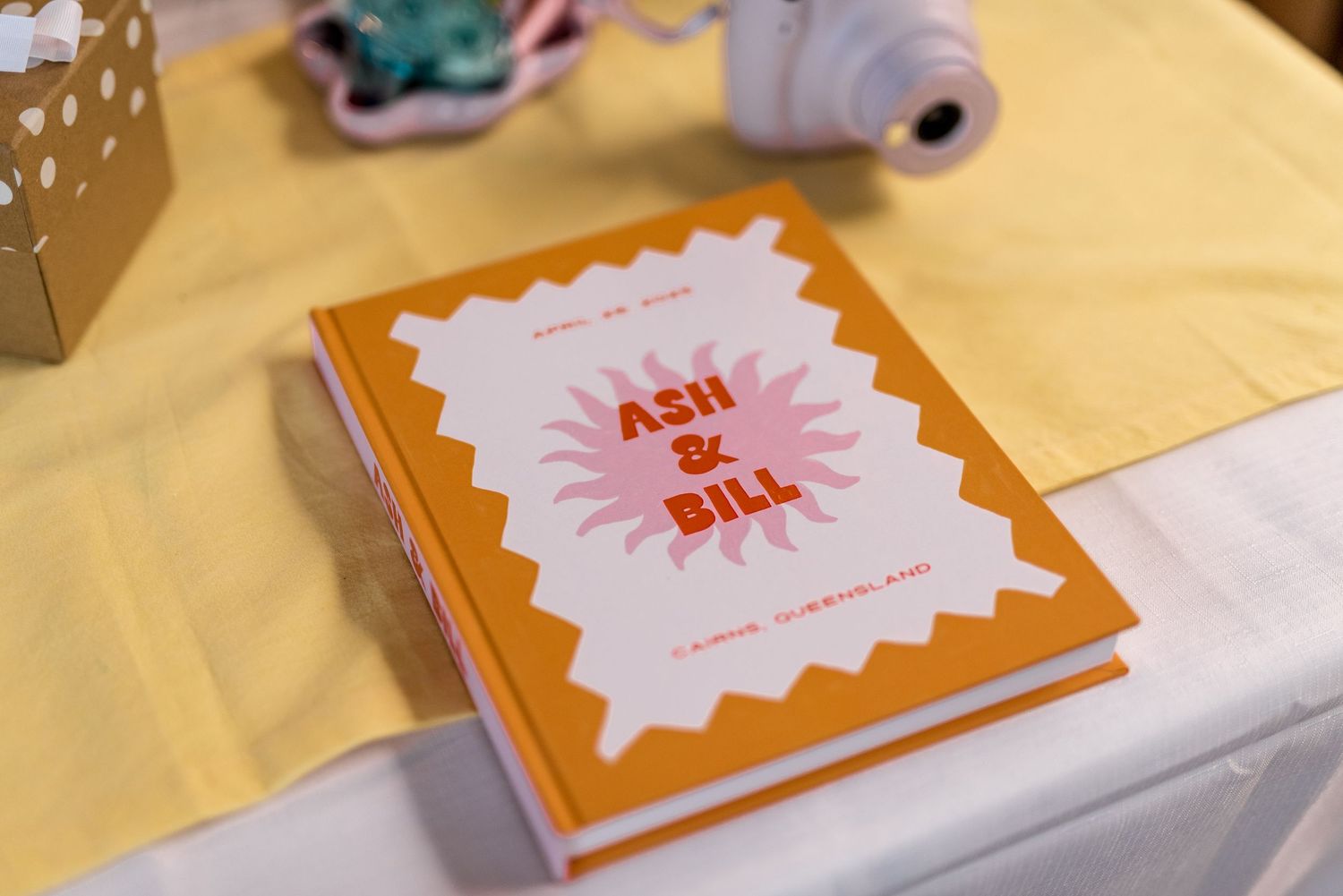 Colorful orange and pink wedding guest book for Ash and Bill displayed on a yellow tablecloth.