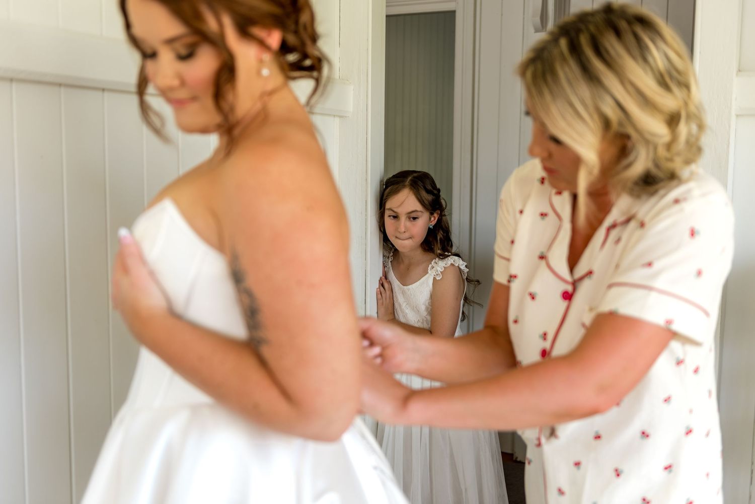 Bride getting wedding dress fastened by blonde woman while young flower girl watches in doorway.