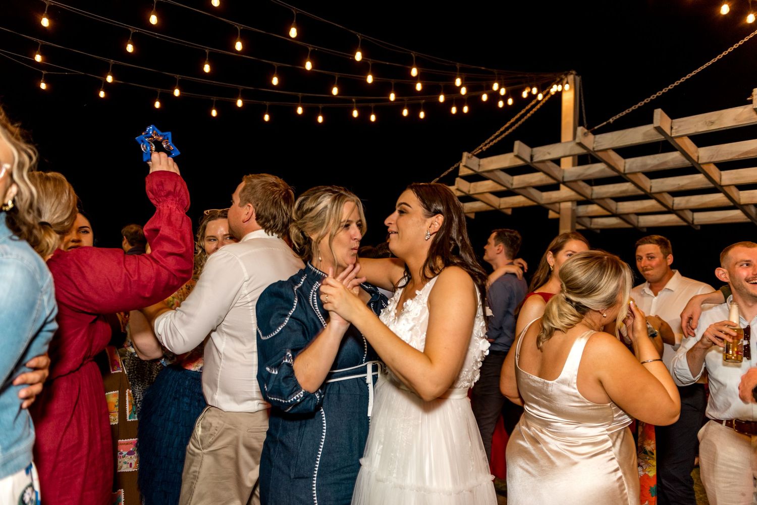 Bride dancing with guests under string lights at outdoor evening wedding reception.