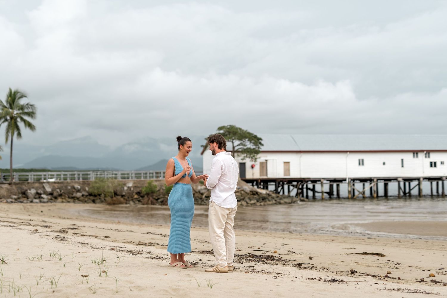 Happy couple sharing engagement moment on sandy beach with white pier building in background.