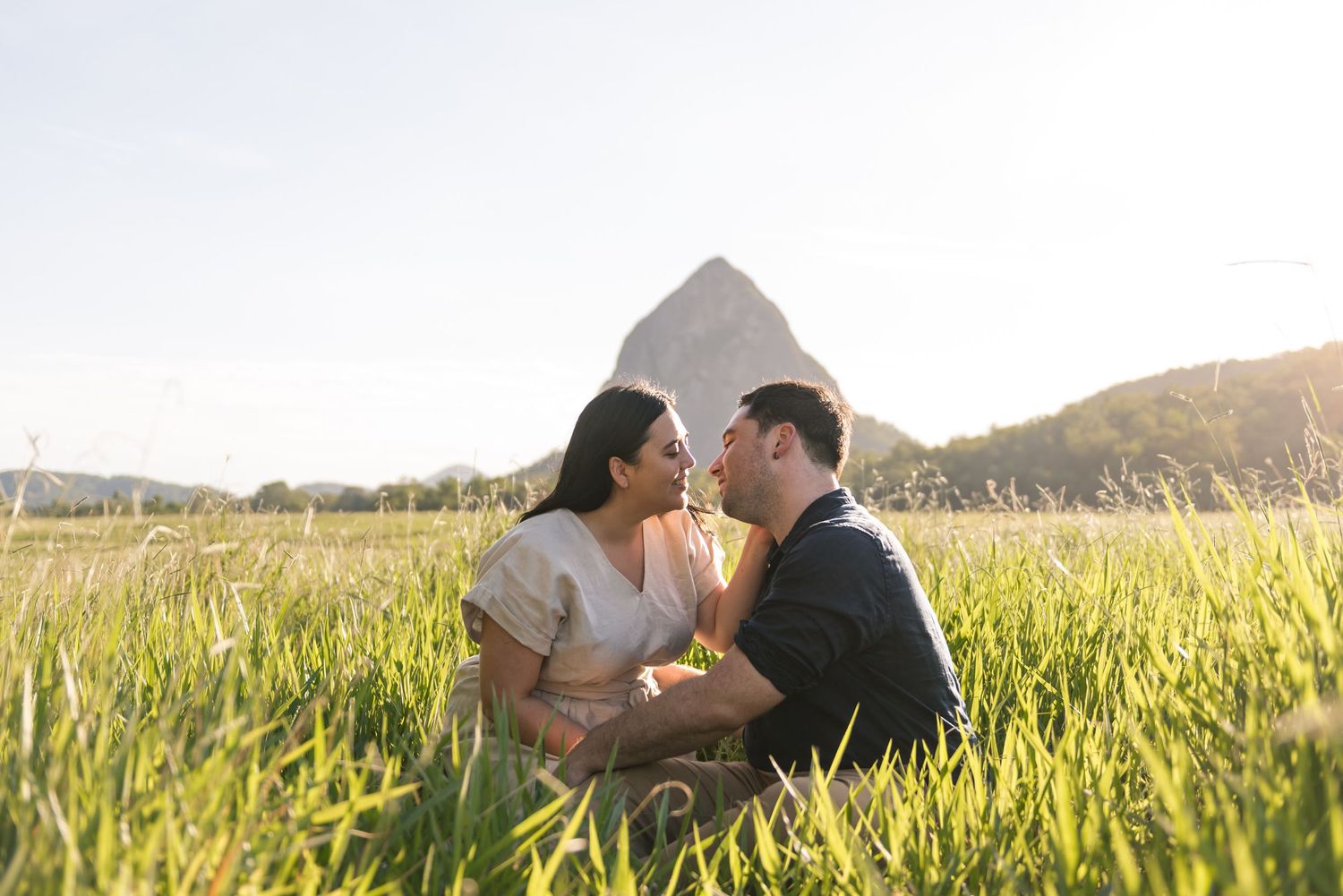 Couple sharing a tender moment sitting in tall green grass with a mountain peak in golden sunlight.