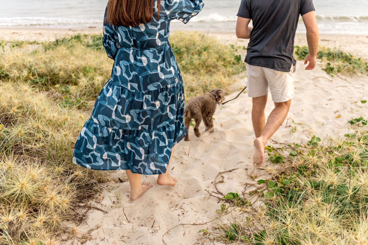 Barefoot couple walking dog on sandy beach path, woman in blue floral dress, man in gray shirt and shorts.