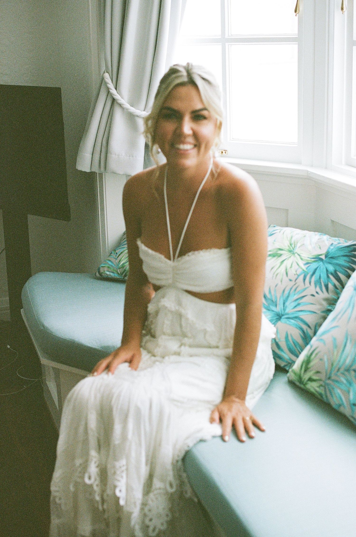 Radiant blonde bride in white strapless lace bridal set sitting on window seat with tropical cushions.