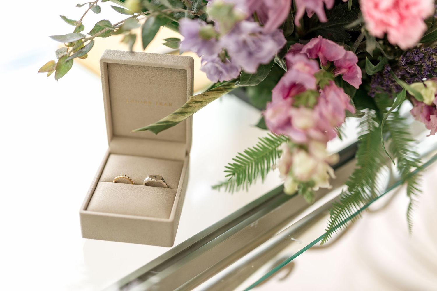 Gold wedding ring in a beige box beside pink and purple floral arrangement.