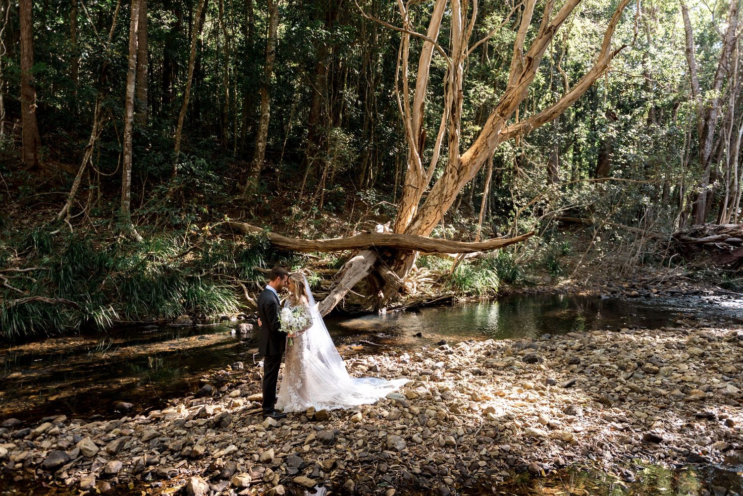 Bride and groom pose together near a creek in a sunlit forest setting.