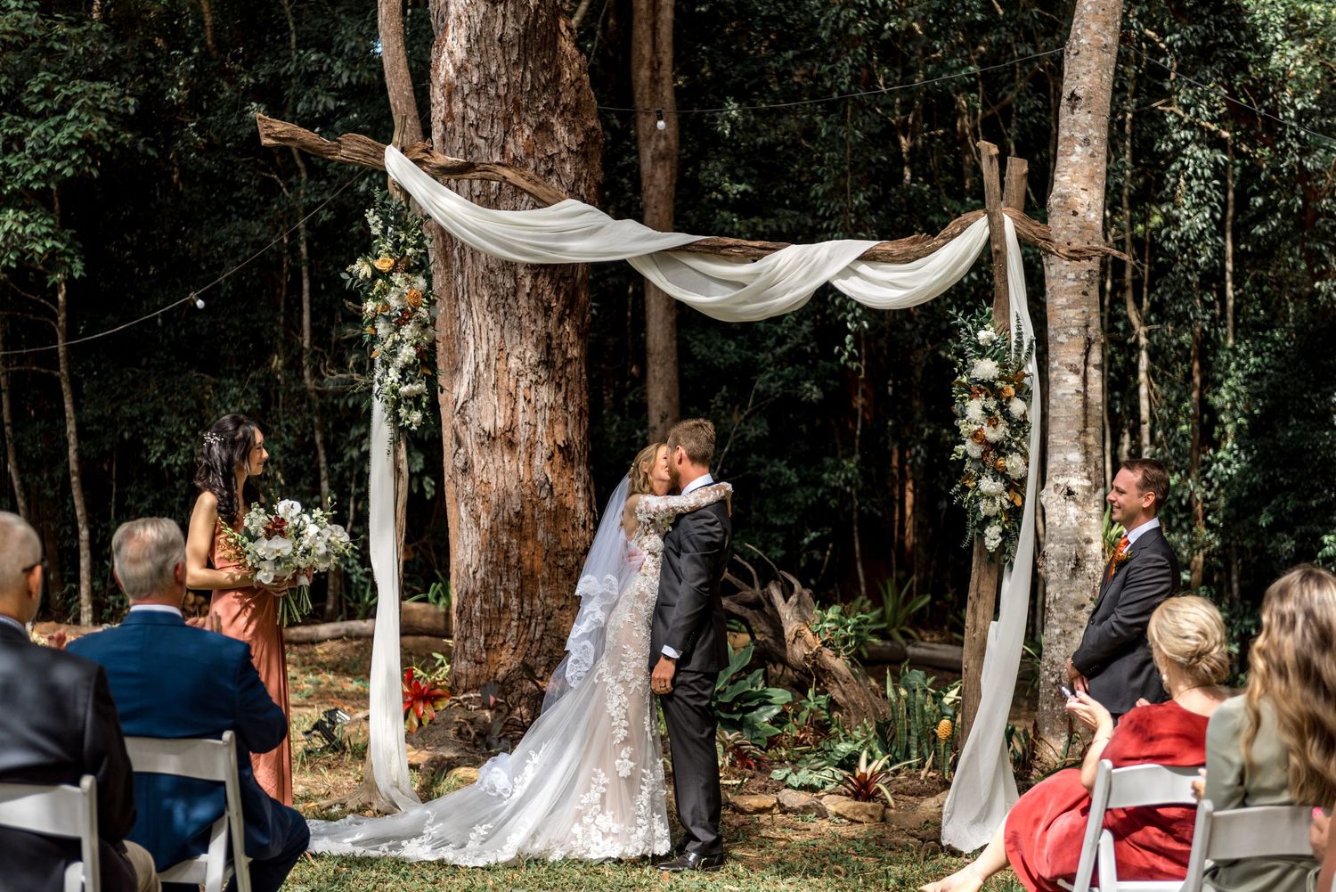Bride and groom share first kiss under draped floral arch in forest wedding.