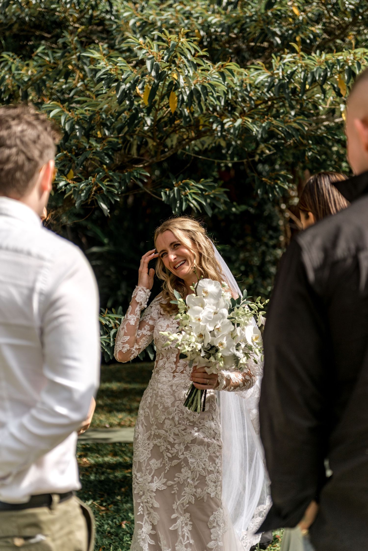 Smiling bride in lace long-sleeve wedding gown holds white bouquet during outdoor ceremony.