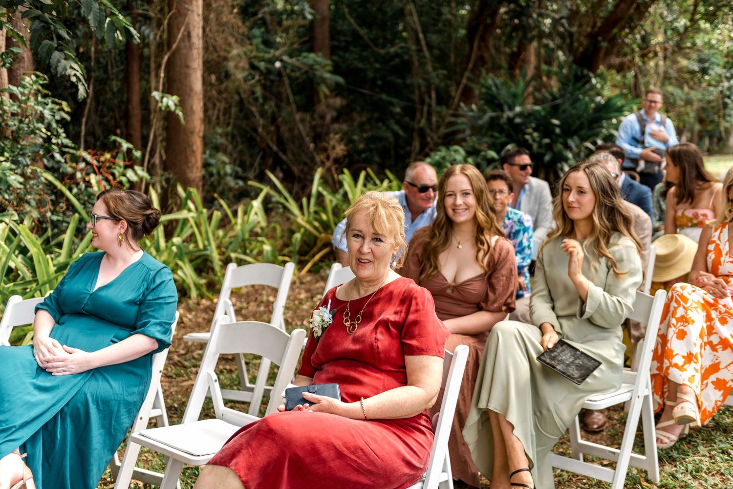 Wedding guests seated in white chairs outdoors, smiling and enjoying the forest ceremony setting.