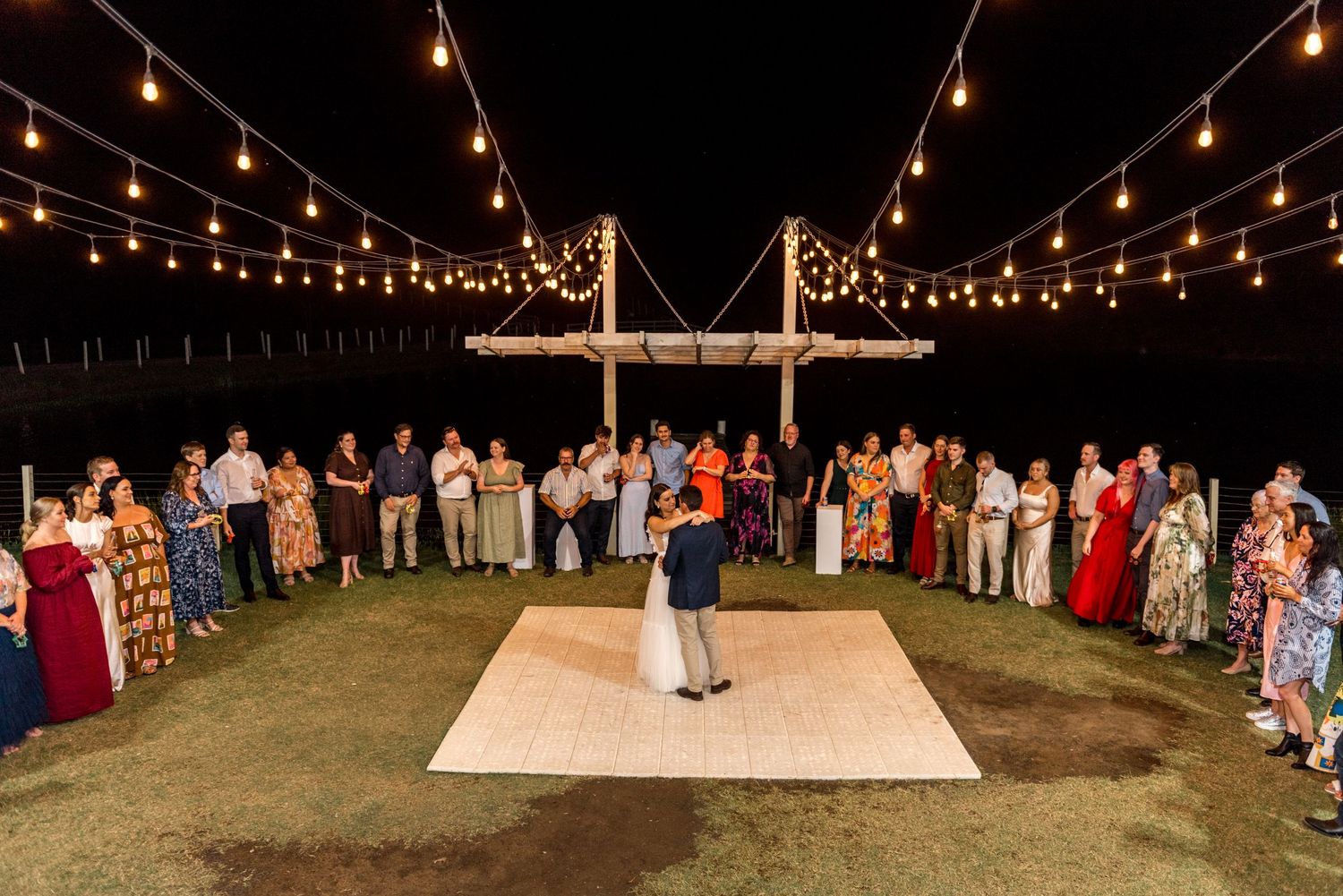 Couple sharing first dance on outdoor dance floor under string lights at night.