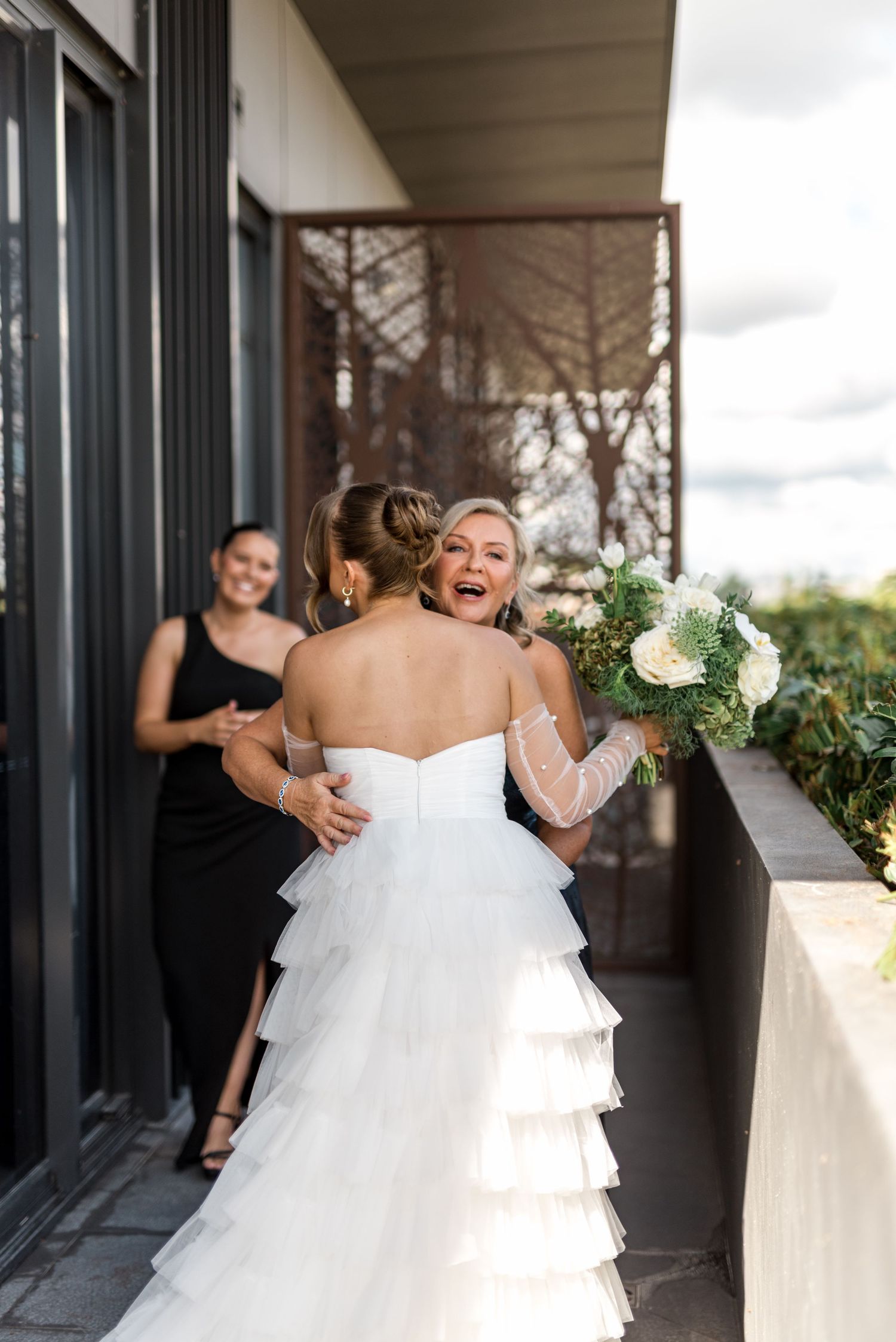 Bride in layered tulle gown embracing emotional guest on balcony while bridesmaid smiles in background.