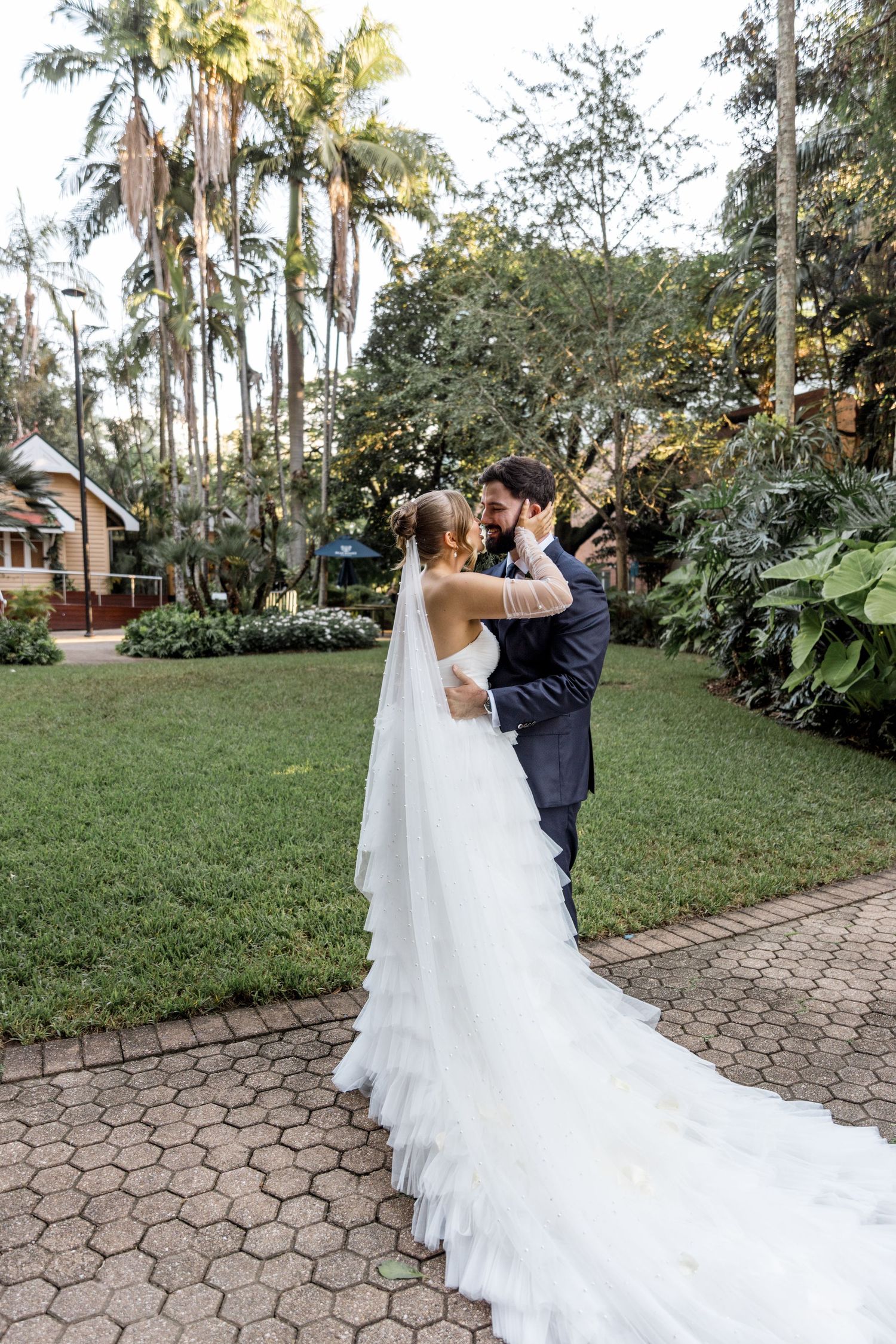 Bride and groom share a romantic embrace in a lush garden setting, bride wearing a tiered white gown with long veil.