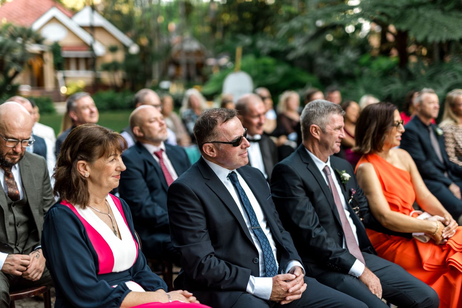 Wedding guests in formal attire seated outdoors smiling during garden wedding ceremony.