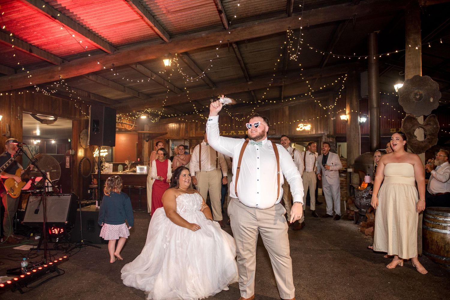 Groom tosses garter while bride sits at rustic barn wedding reception with string lights.