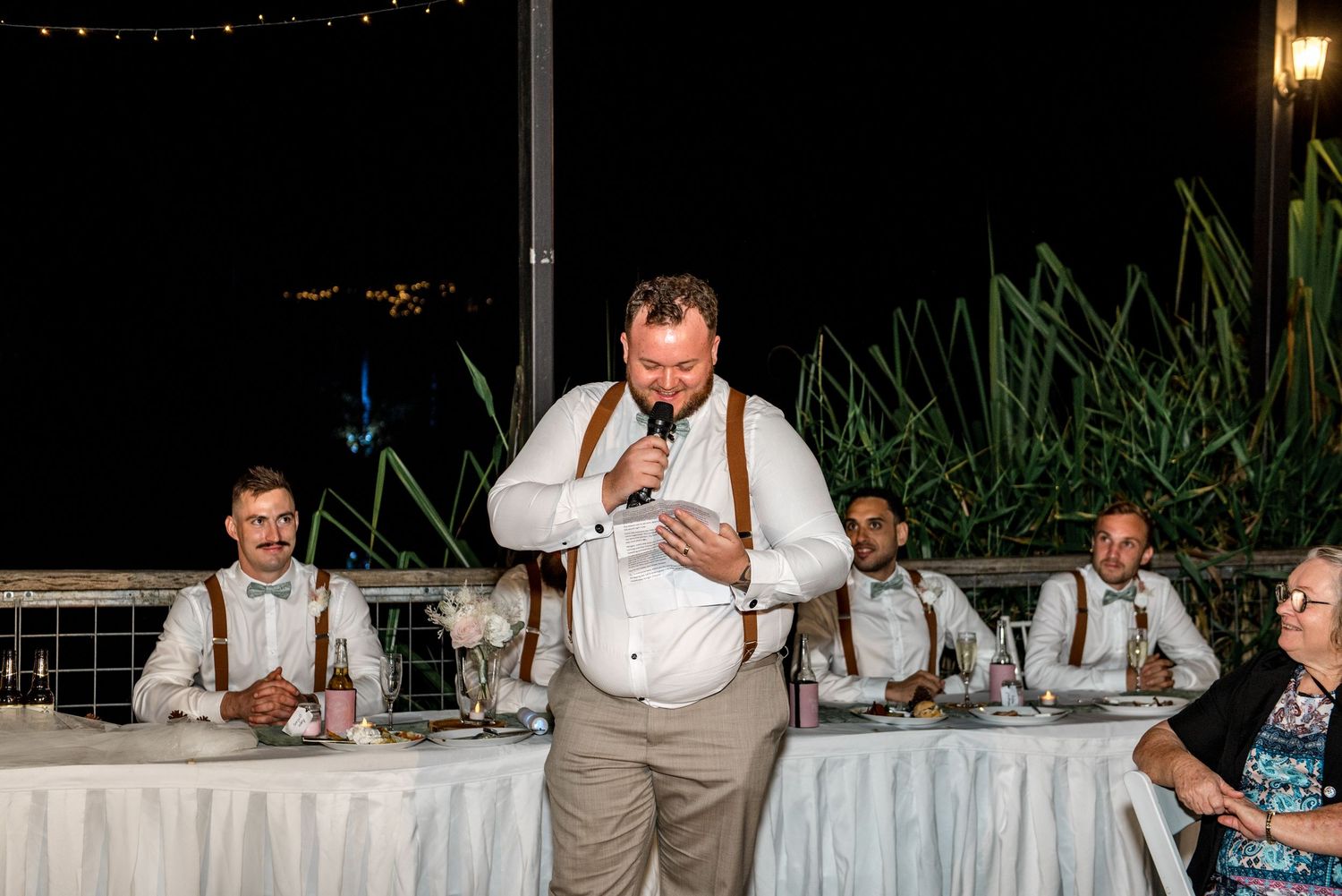 Groomsman delivers emotional wedding speech at reception with bridal party seated at head table.