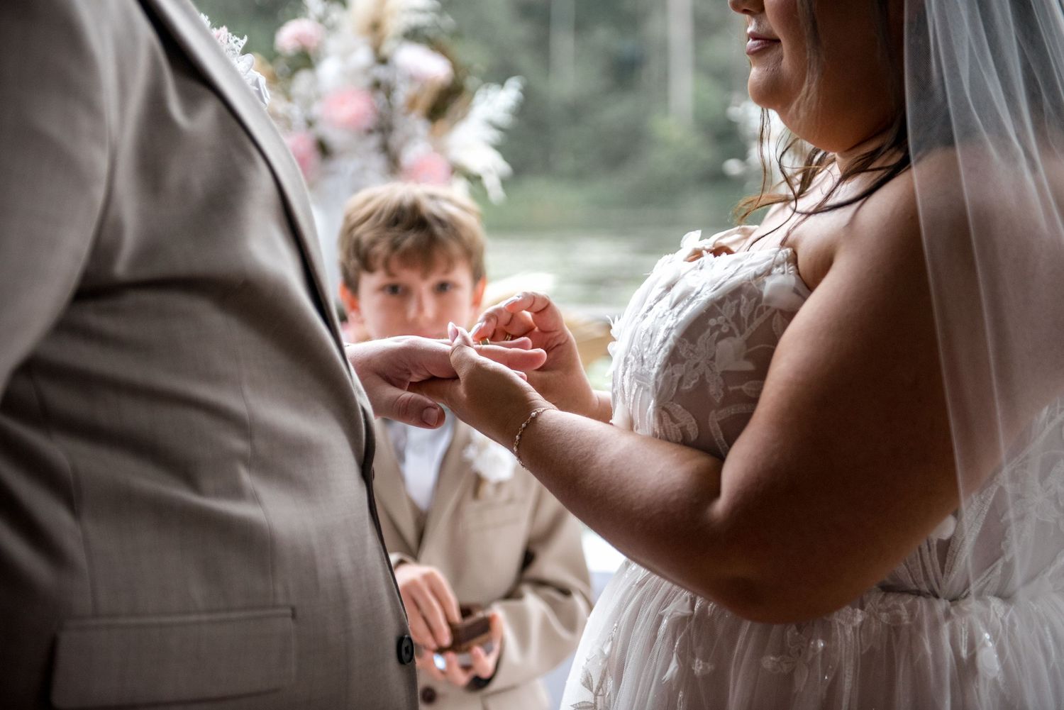 Bride places wedding ring on grooms finger during emotional ring exchange ceremony, young boy visible in background.
