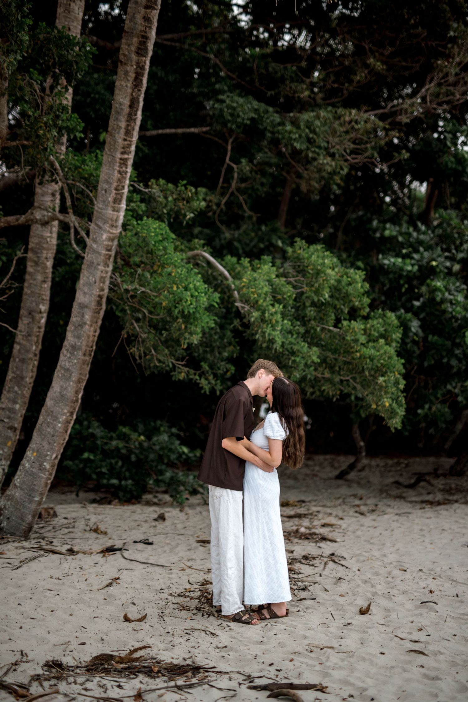 Couple sharing a tender kiss on a sandy beach with tall palm trees in the background.