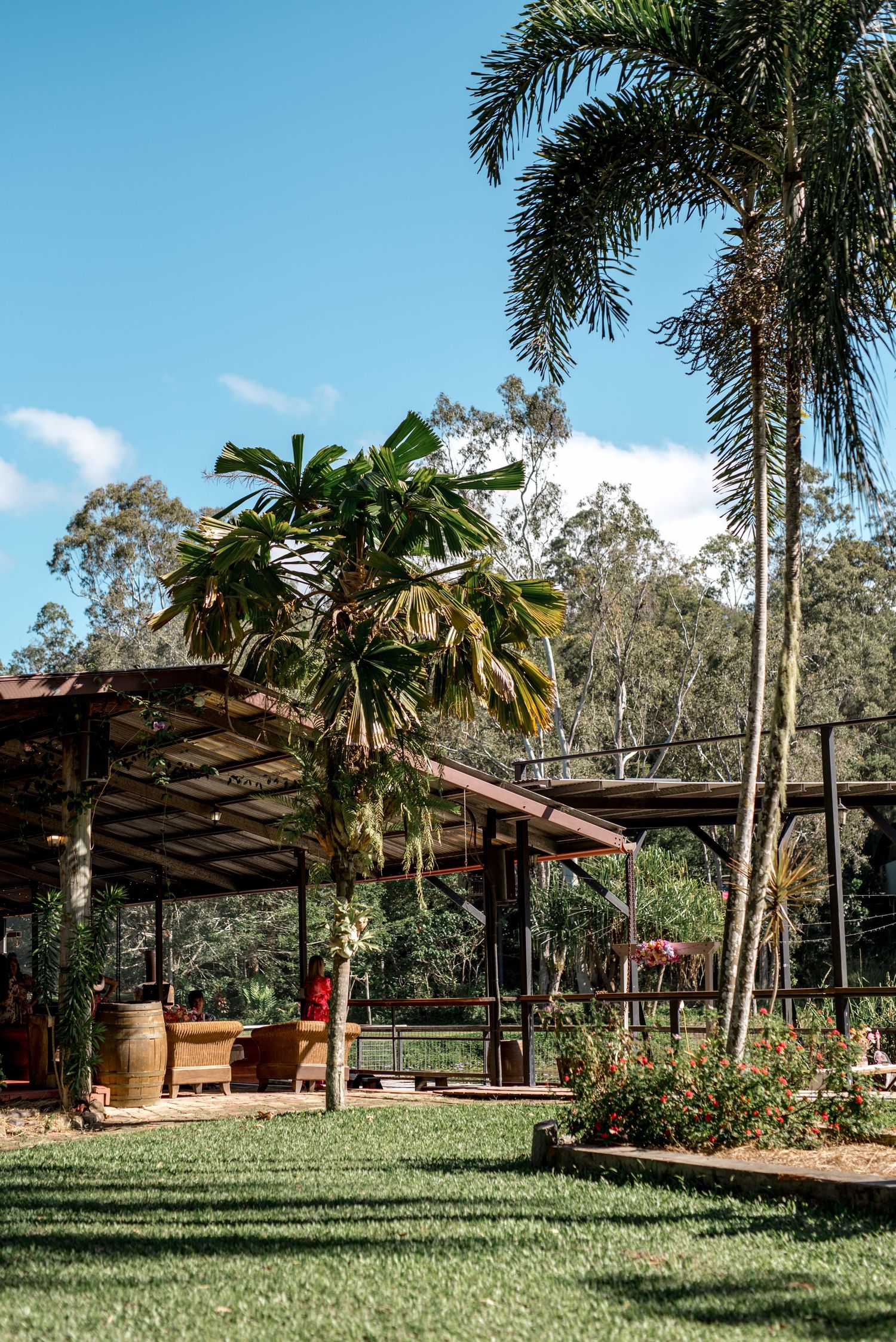 Tropical wedding venue with open-air pavilion, lush palm trees, manicured lawn, and natural bushland backdrop under blue sky.