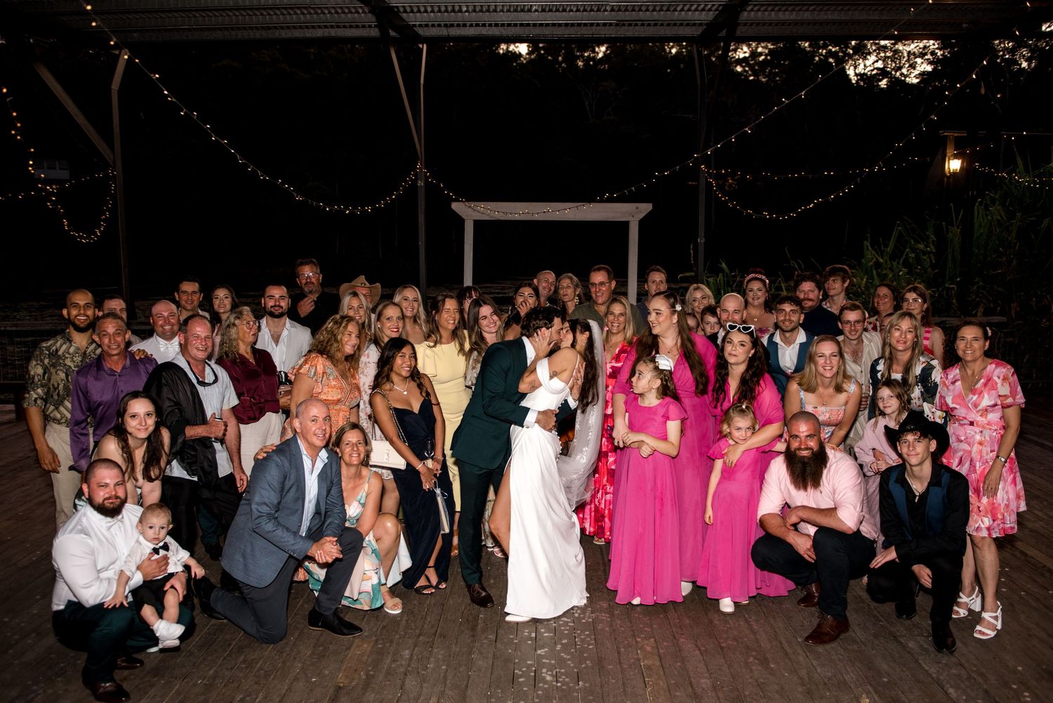 Large wedding group photo with guests gathered outdoors under string lights at a garden venue.