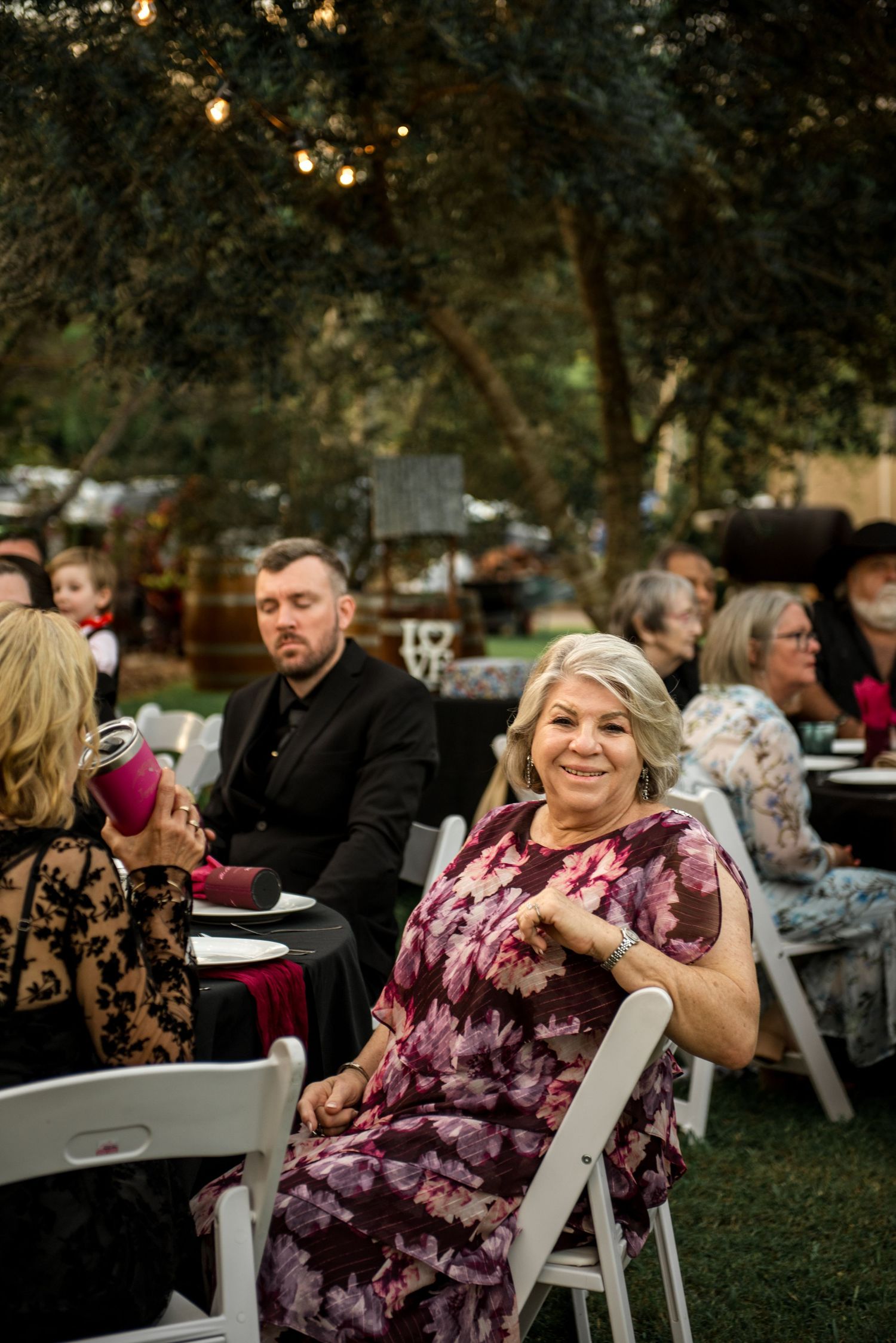 Smiling woman in floral dress seated at outdoor wedding reception with string lights and guests.