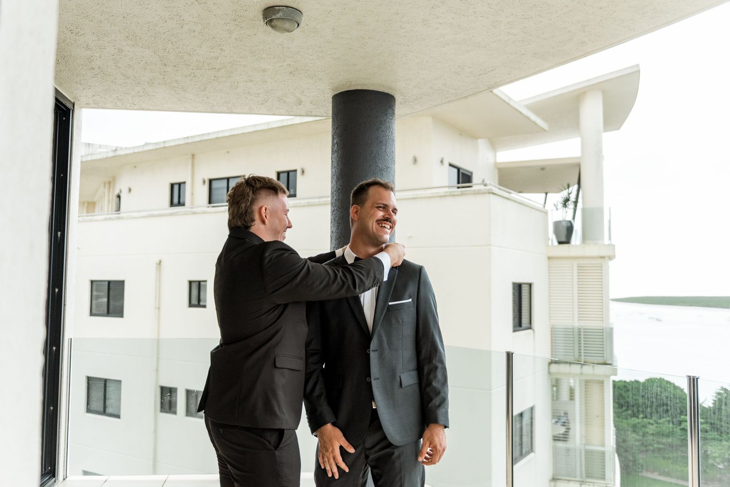 Two men in black suits share a joyful moment on a hotel balcony during wedding preparations.