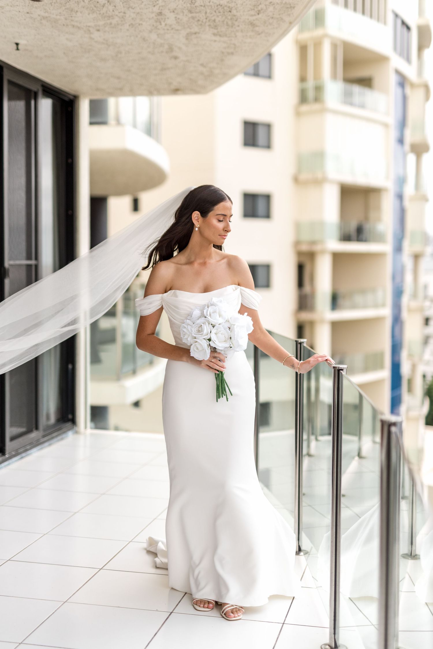 Bride in elegant white off-shoulder gown holding white bouquet on sunny balcony.
