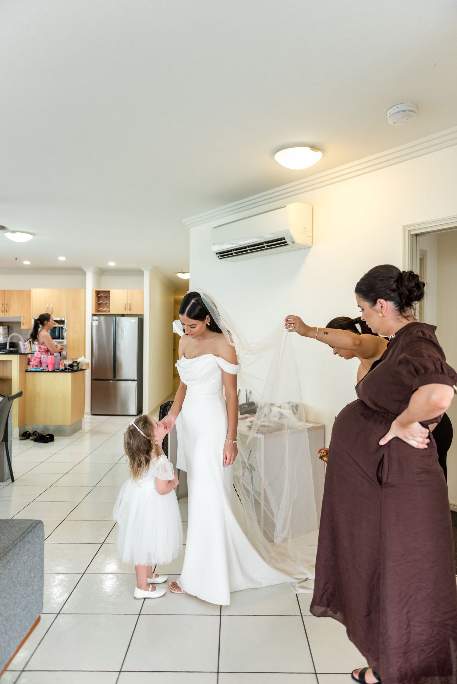 Bride in white gown and veil holds hands with flower girl while assistant arranges train indoors.