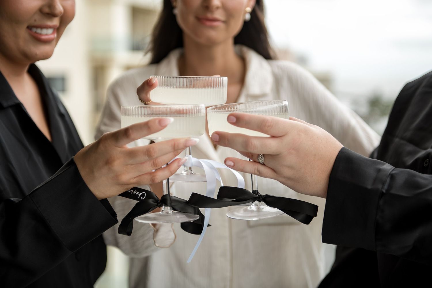 Three women clinking champagne glasses in a celebratory toast during wedding preparations, wearing black and white outfits.