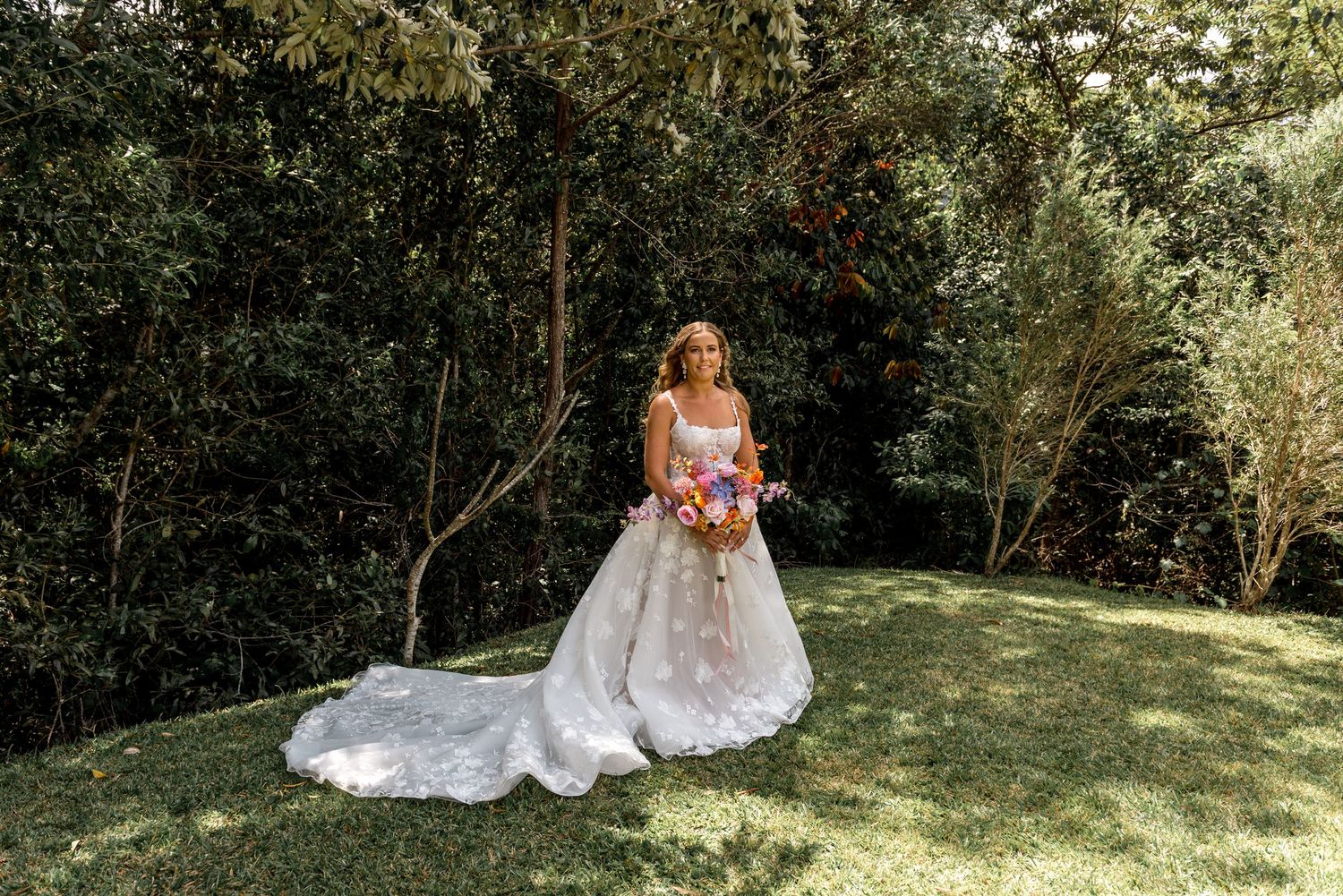 Bride holding colorful bouquet in a floral lace A-line wedding gown standing on a sunlit green lawn.