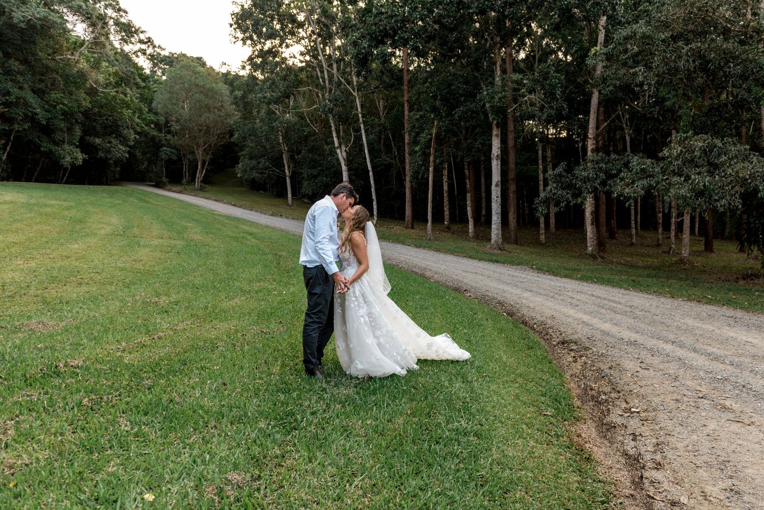 Bride and groom embrace romantically on a green lawn beside a gravel path in a lush forest setting.