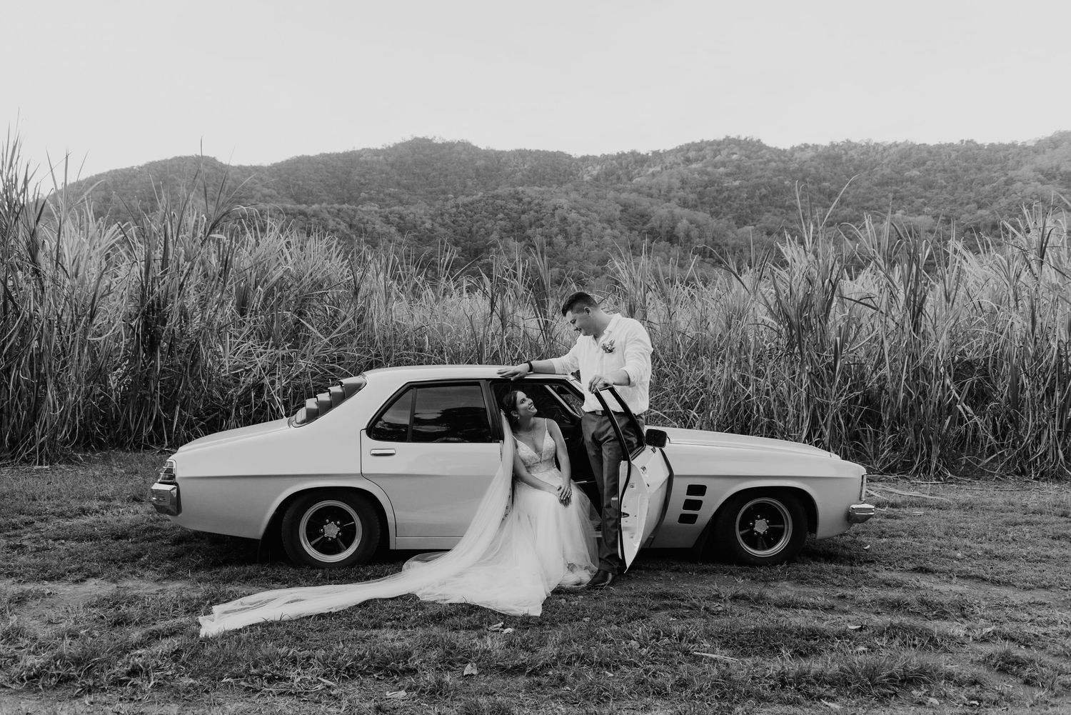 Black and white photo of bride and groom posing with a vintage classic car outdoors.