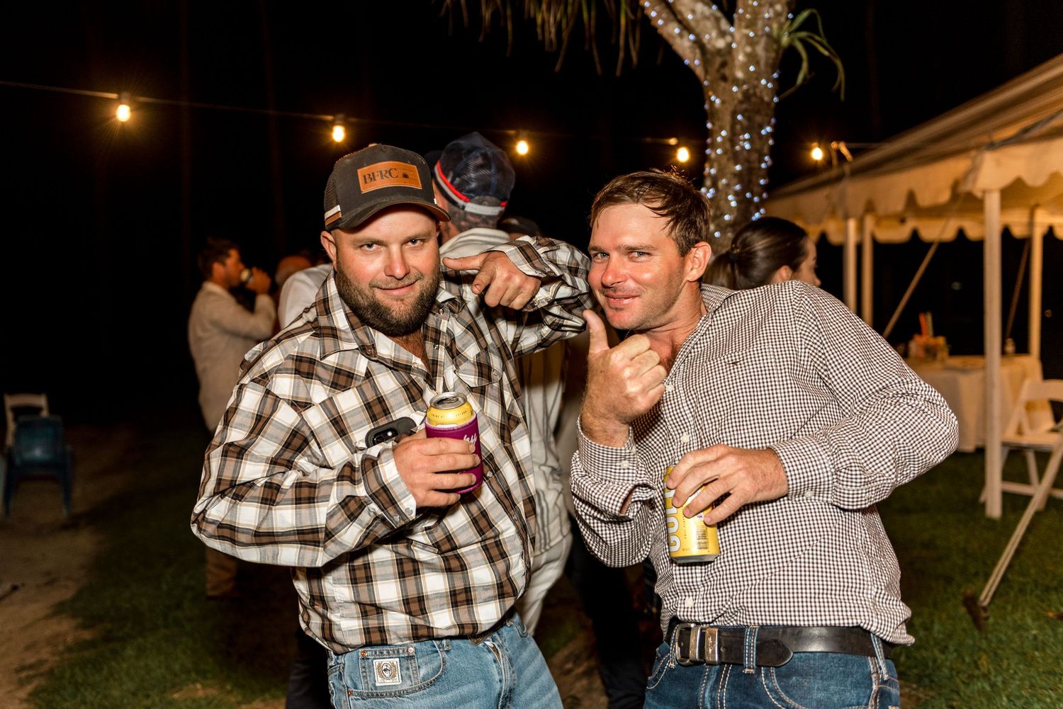 Two men smiling and posing with drinks at an outdoor evening party, wearing flannel and casual shirts.