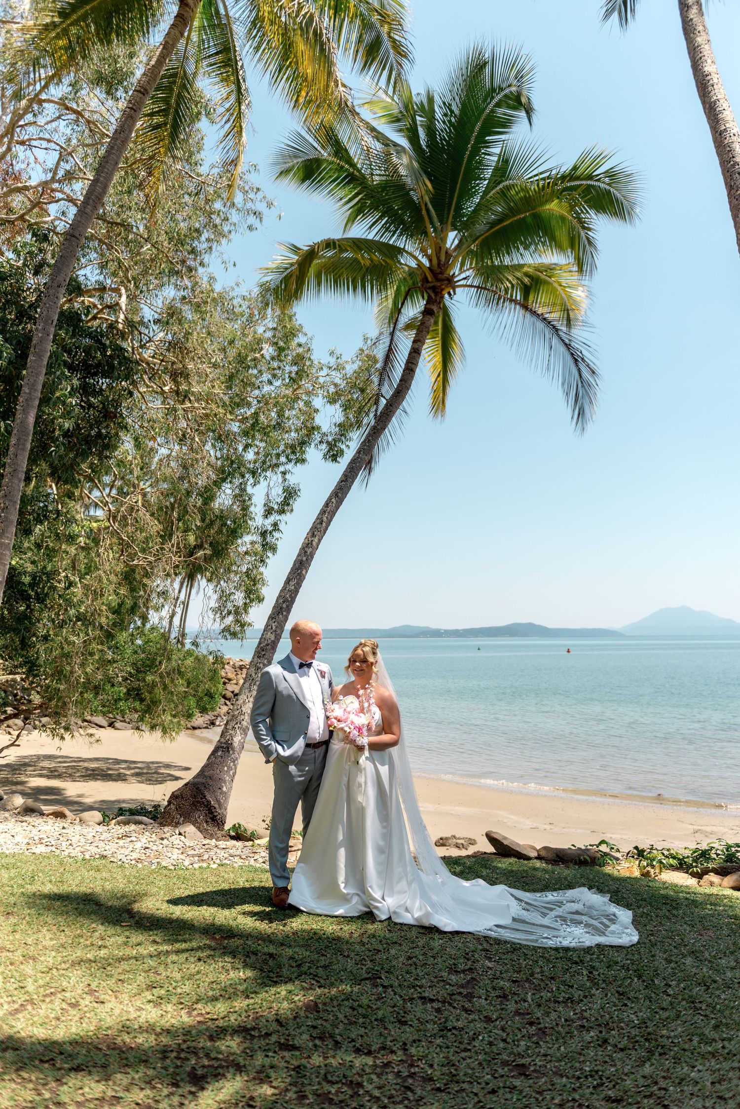 Bride and groom pose together on a sunny tropical beach surrounded by palm trees and calm turquoise waters.