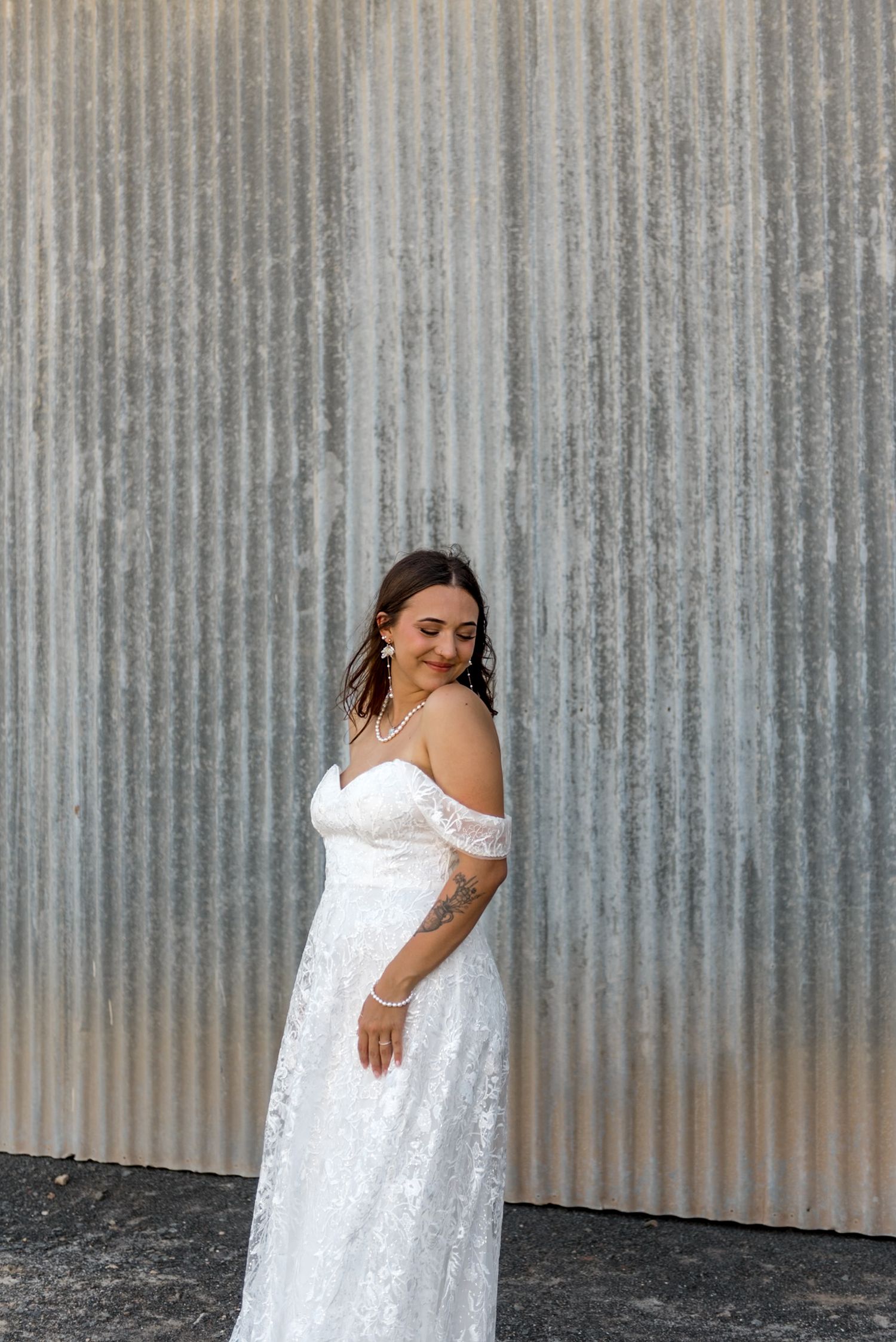 Radiant bride in off-shoulder lace wedding dress smiles softly while posing against corrugated metal wall.