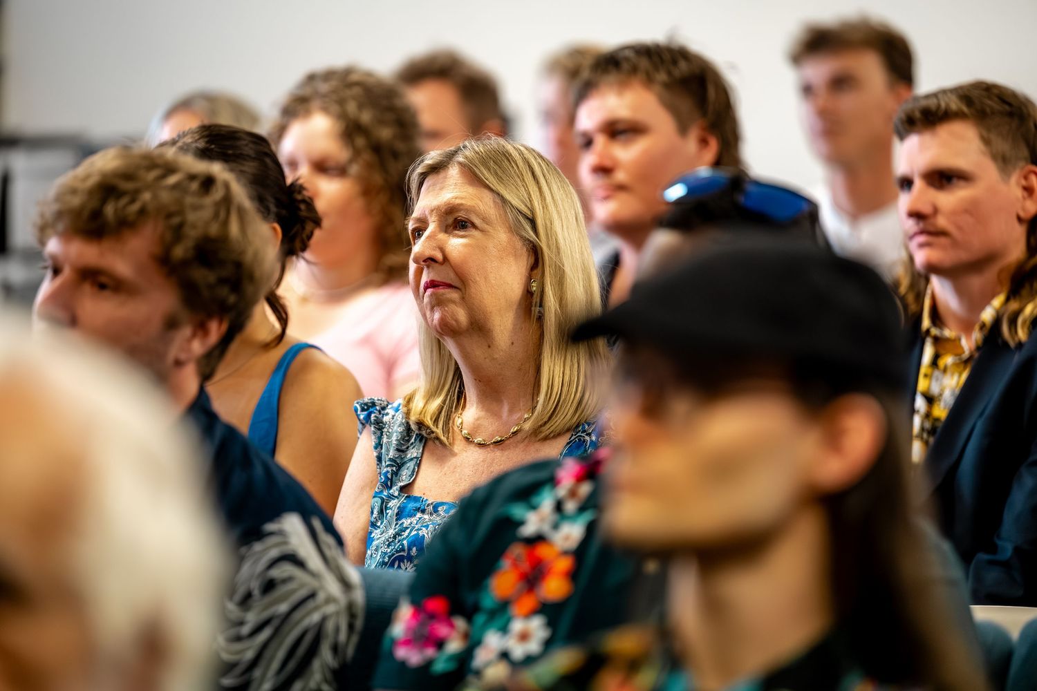 Blonde woman in floral blue dress watches attentively among seated wedding ceremony guests in a crowded venue.