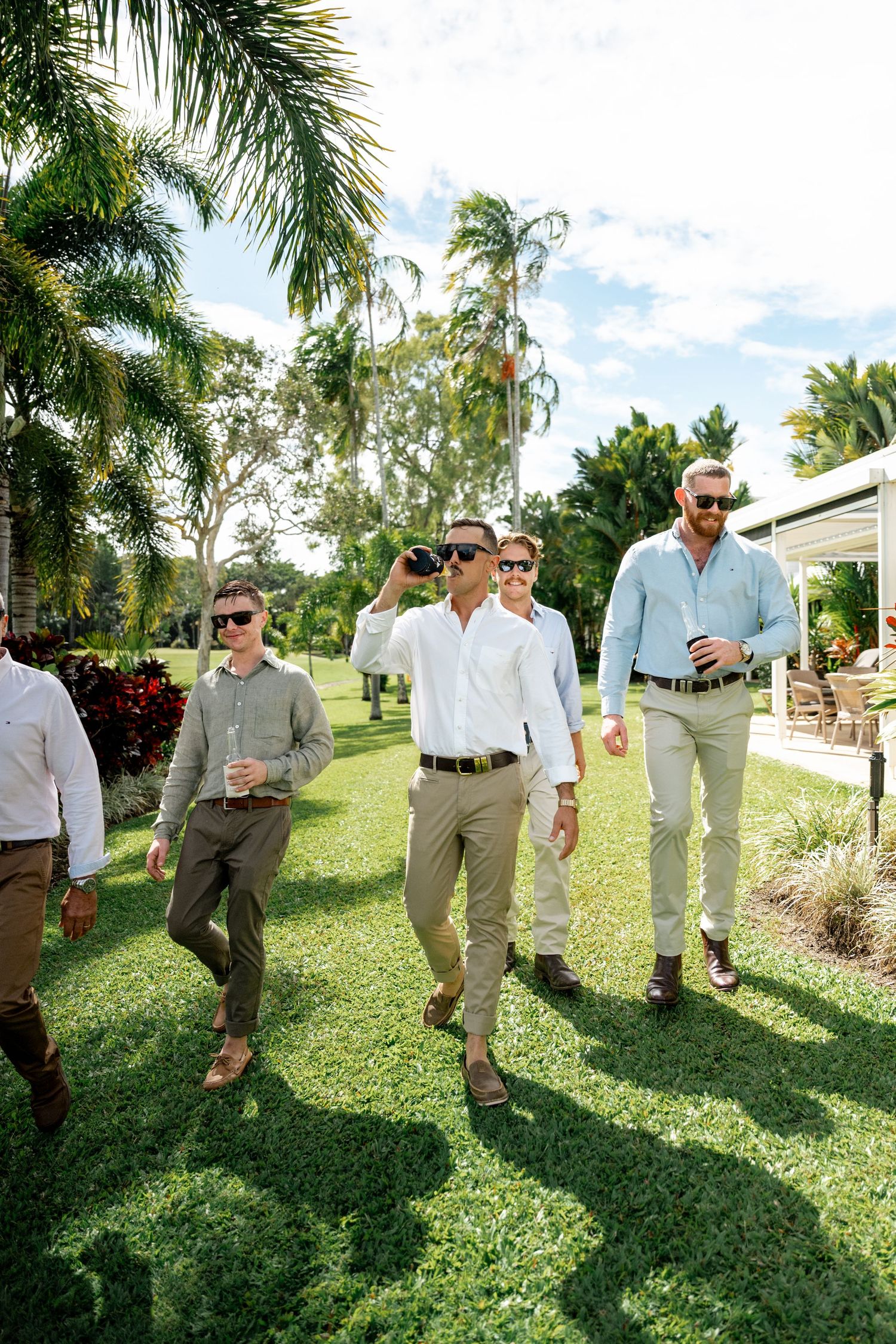 Groomsmen in white shirts and khaki pants walking through tropical garden holding drinks on wedding day.