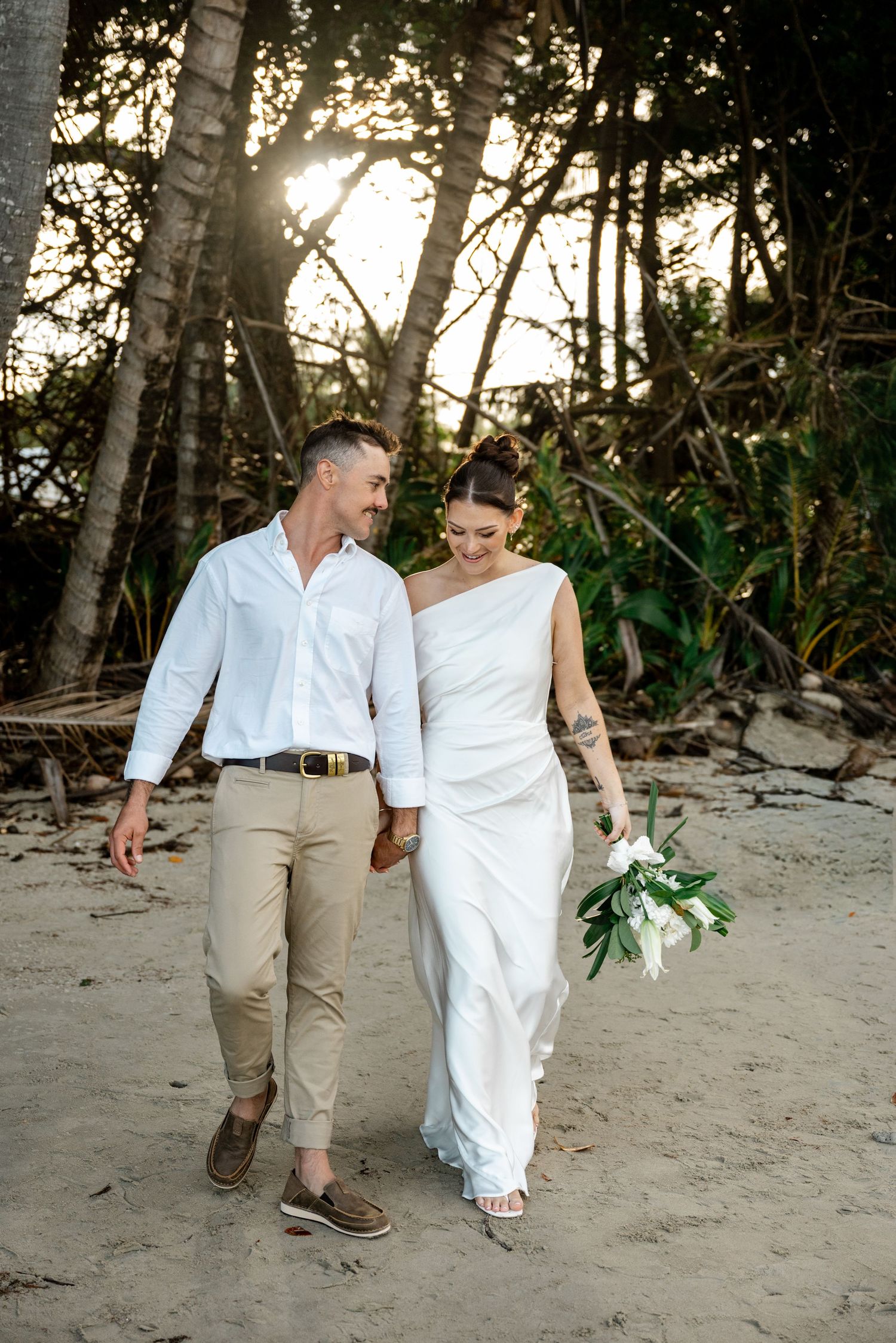Smiling bride and groom stroll along a sandy tropical beach at sunset with white floral bouquet at Four Mile Beach, Port Douglas.