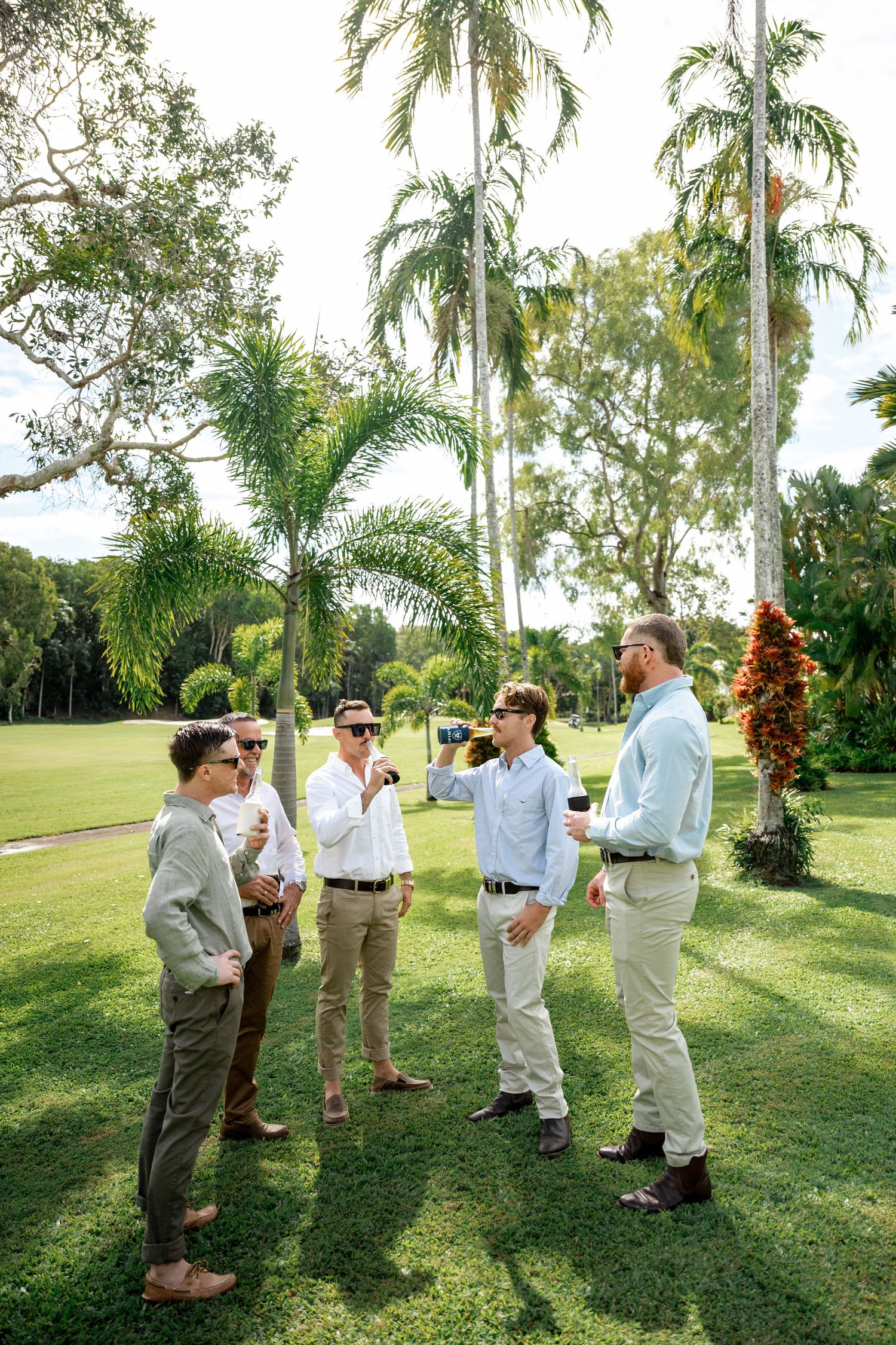 Five groomsmen in casual attire socializing on a green lawn surrounded by tall tropical palm trees.