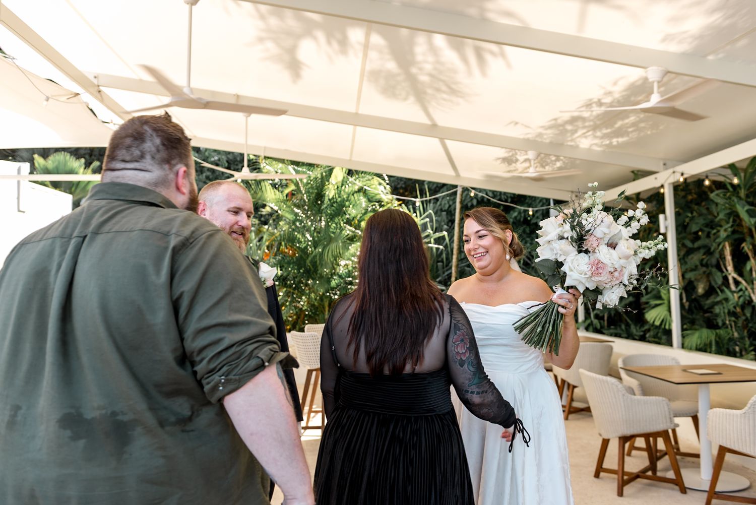Bride smiles holding white floral bouquet while greeting guests at outdoor wedding reception.