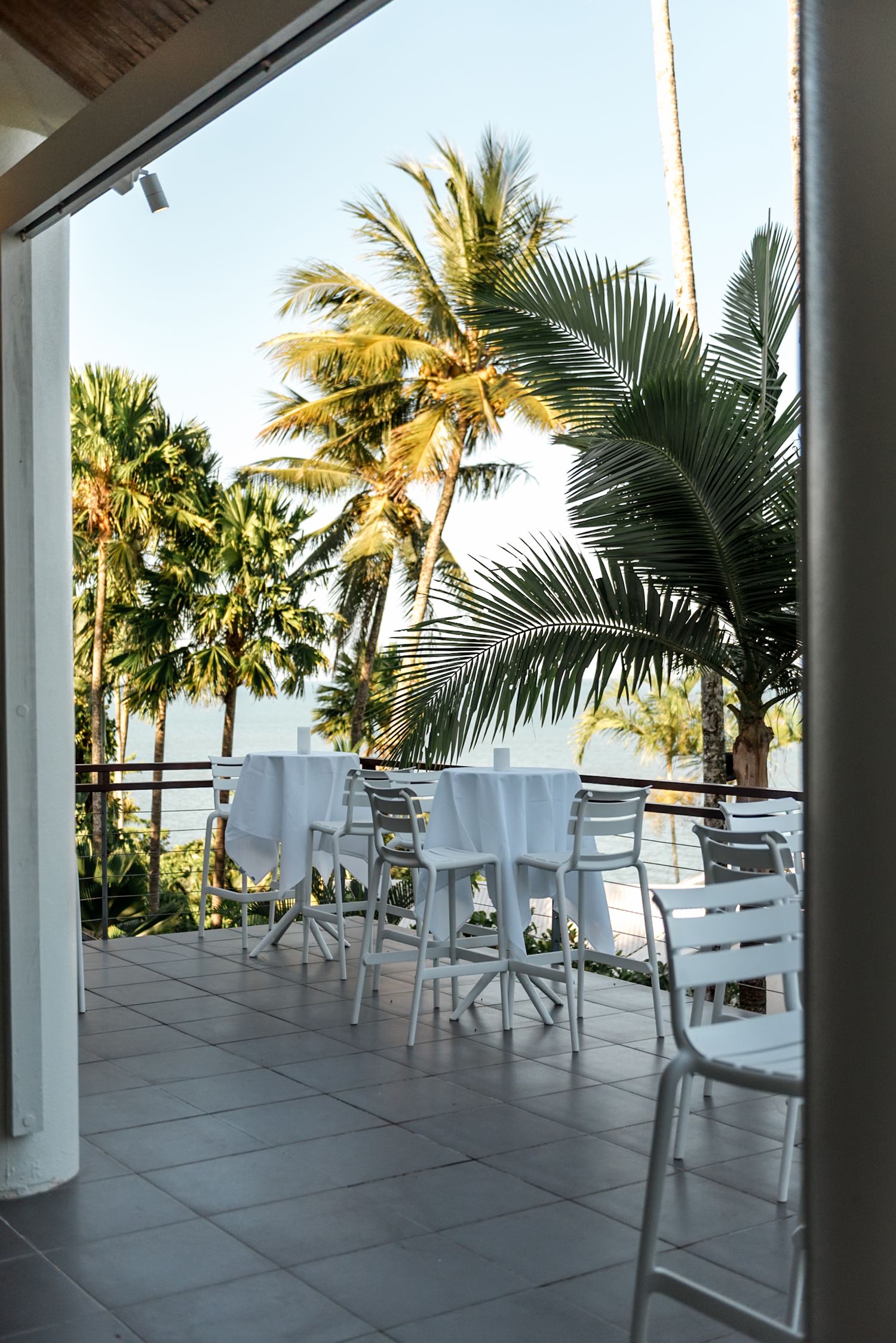 Tropical wedding venue outdoor terrace with white tables, chairs, and palm trees overlooking the ocean.