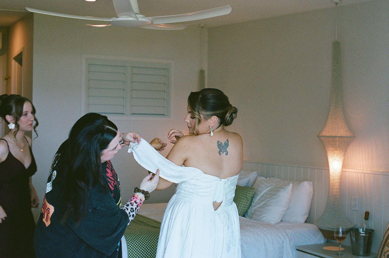 Bride in off-shoulder white gown gets dressed with help from bridesmaids in a sunlit room.