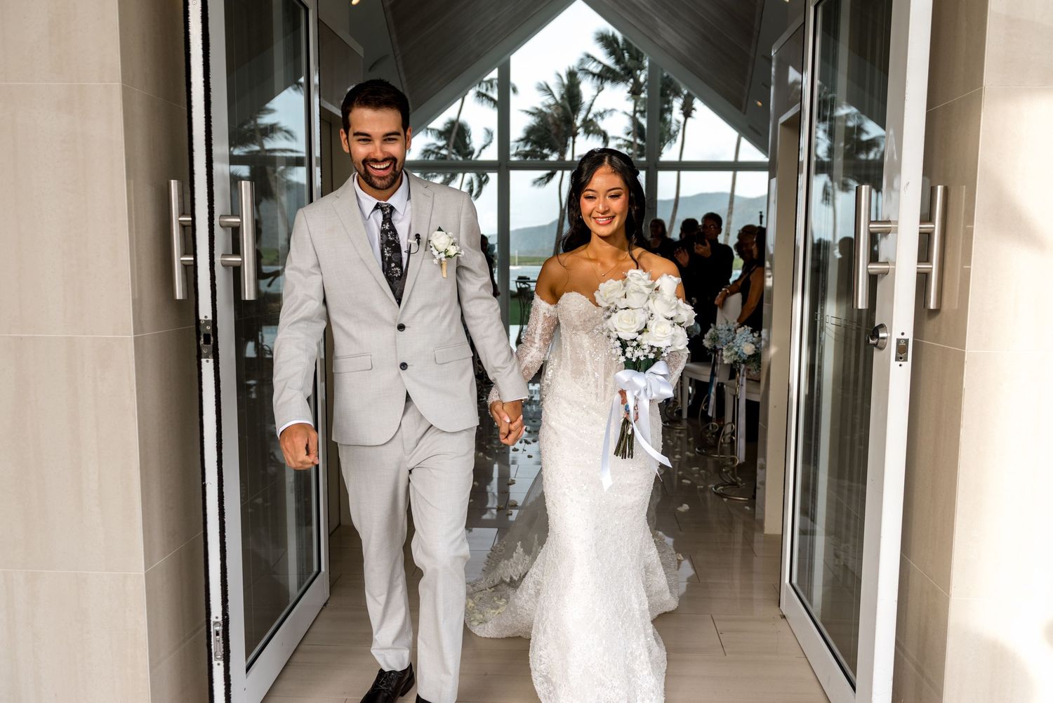 Joyful newlyweds exit stunning glass chapel hand-in-hand with tropical ocean views in background.