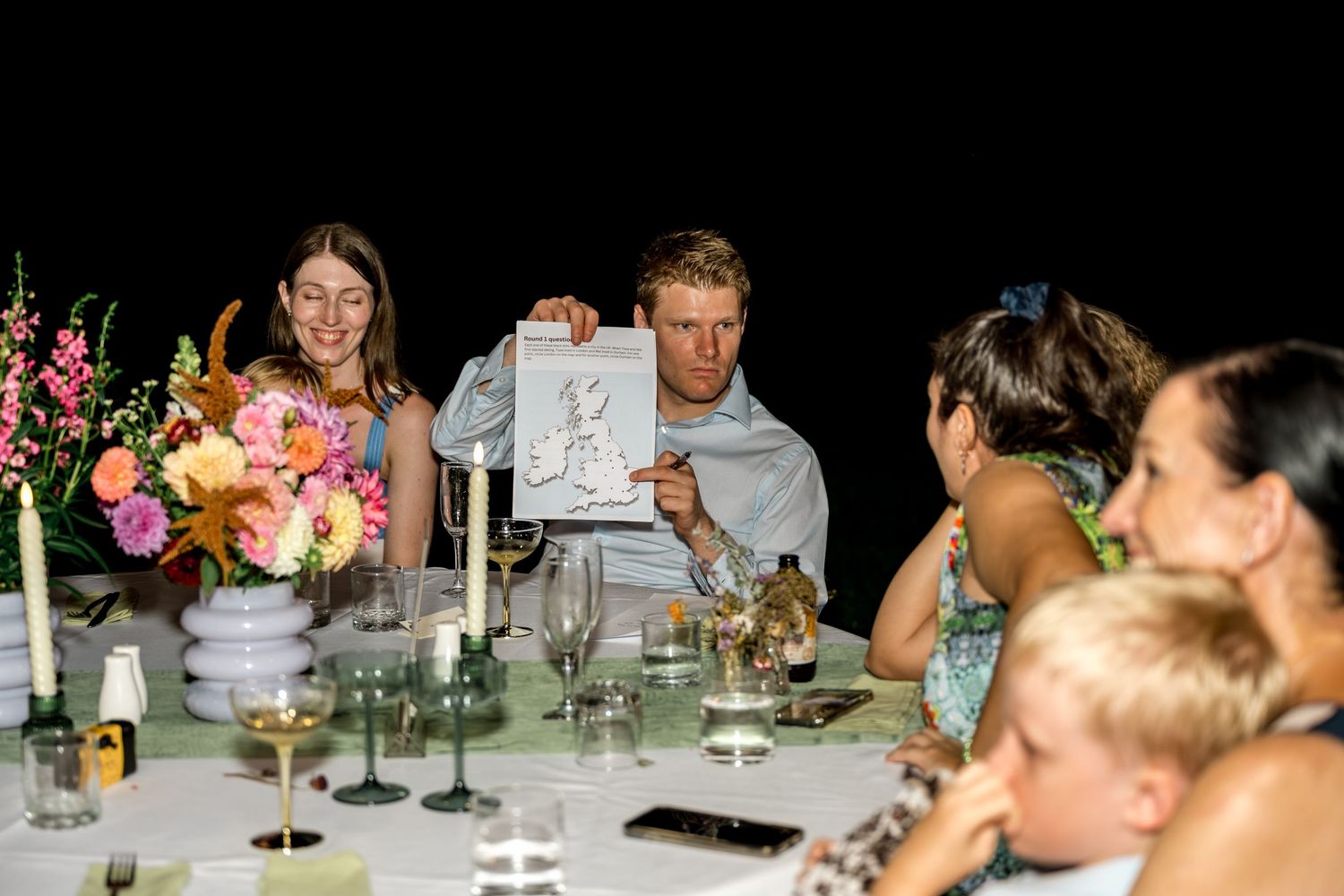 Man proudly displaying a drawing to guests at a floral-decorated dinner table during an event.