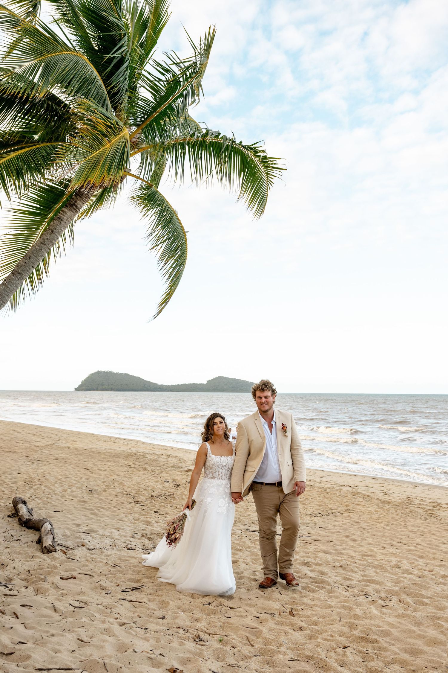 Bride and groom walk hand-in-hand along a tropical beach under swaying palm trees with ocean views.