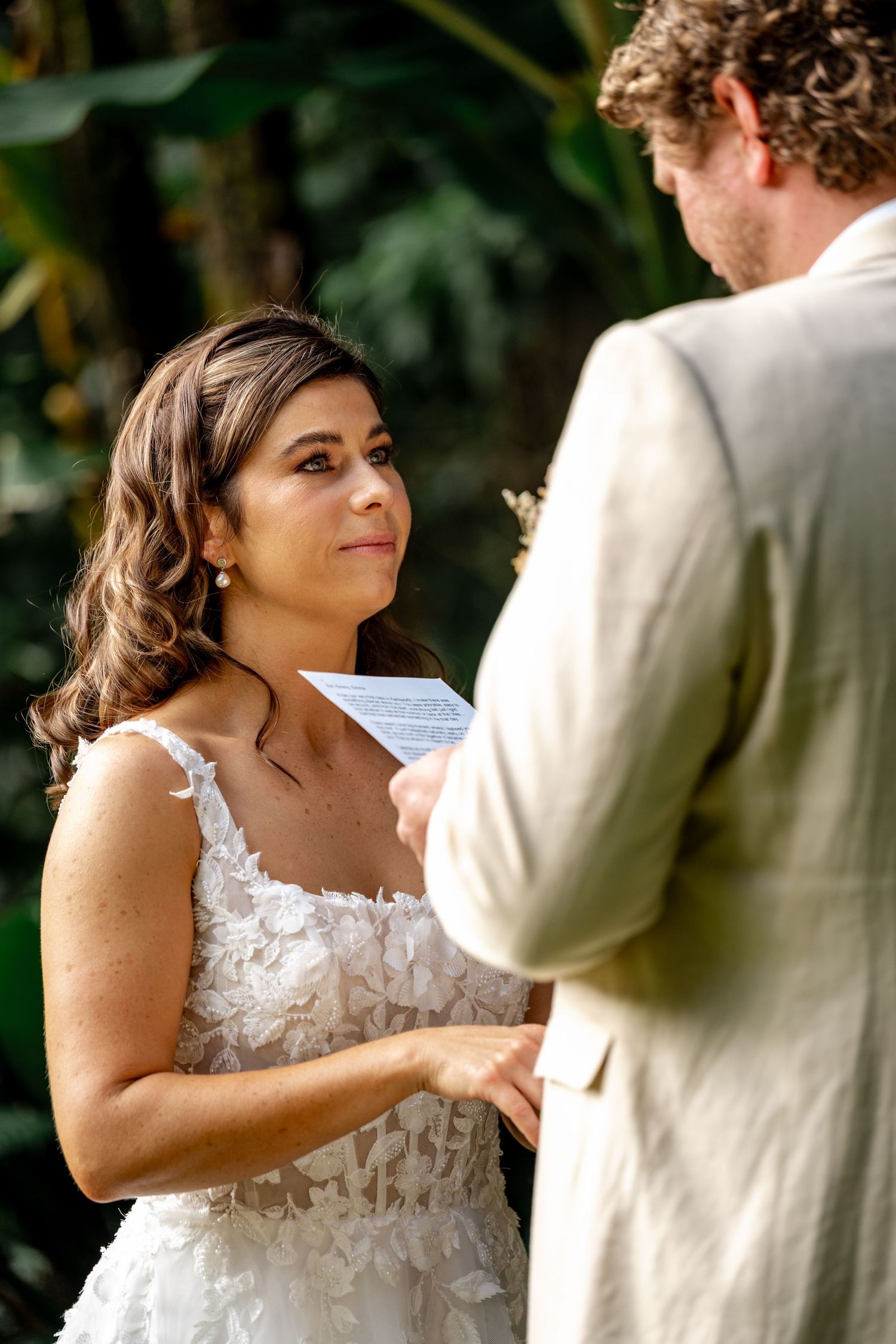 Emotional bride reads wedding vows while holding grooms hands during outdoor tropical garden ceremony.