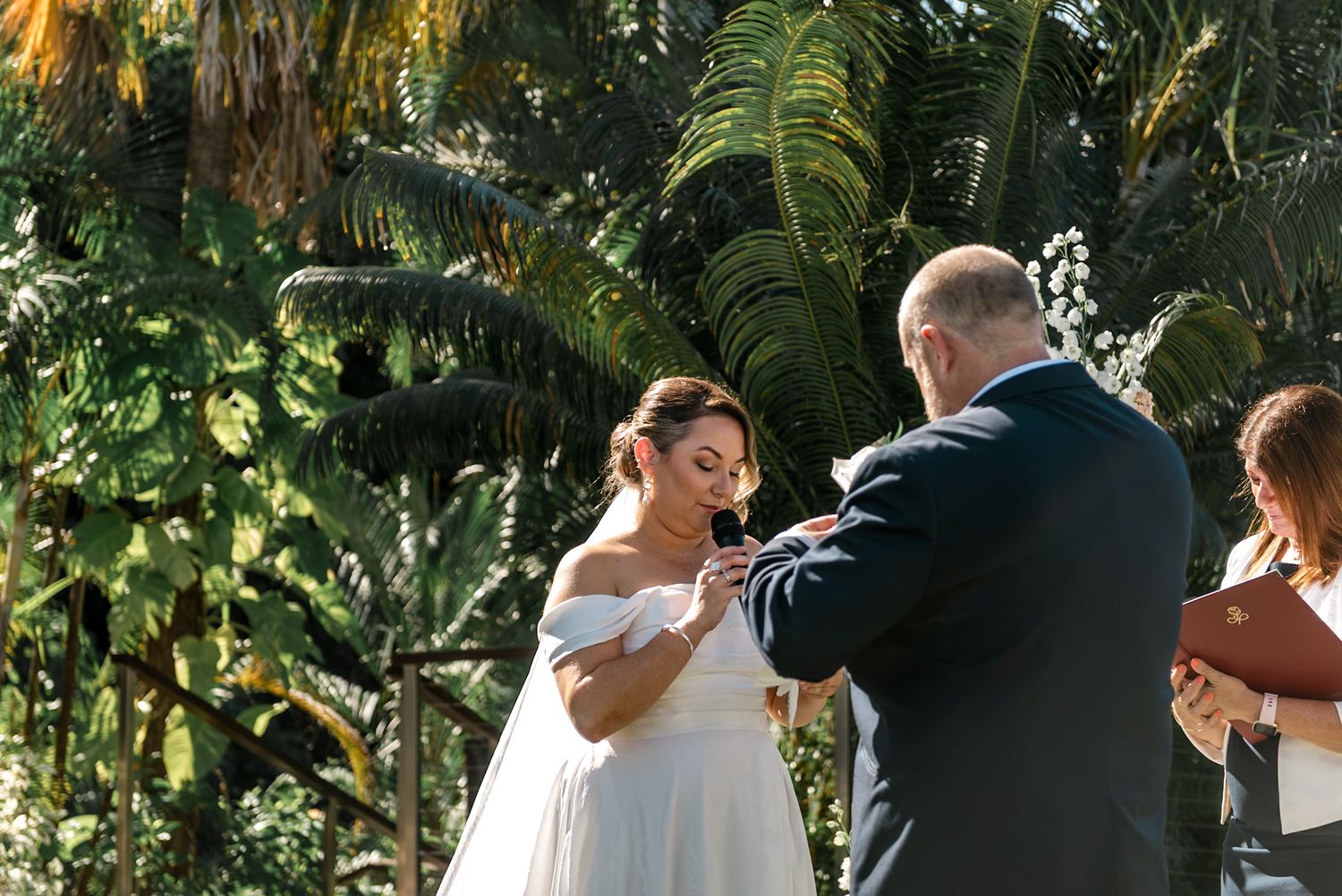 Bride in off-shoulder white gown exchanges rings with groom during outdoor wedding ceremony among lush palms.