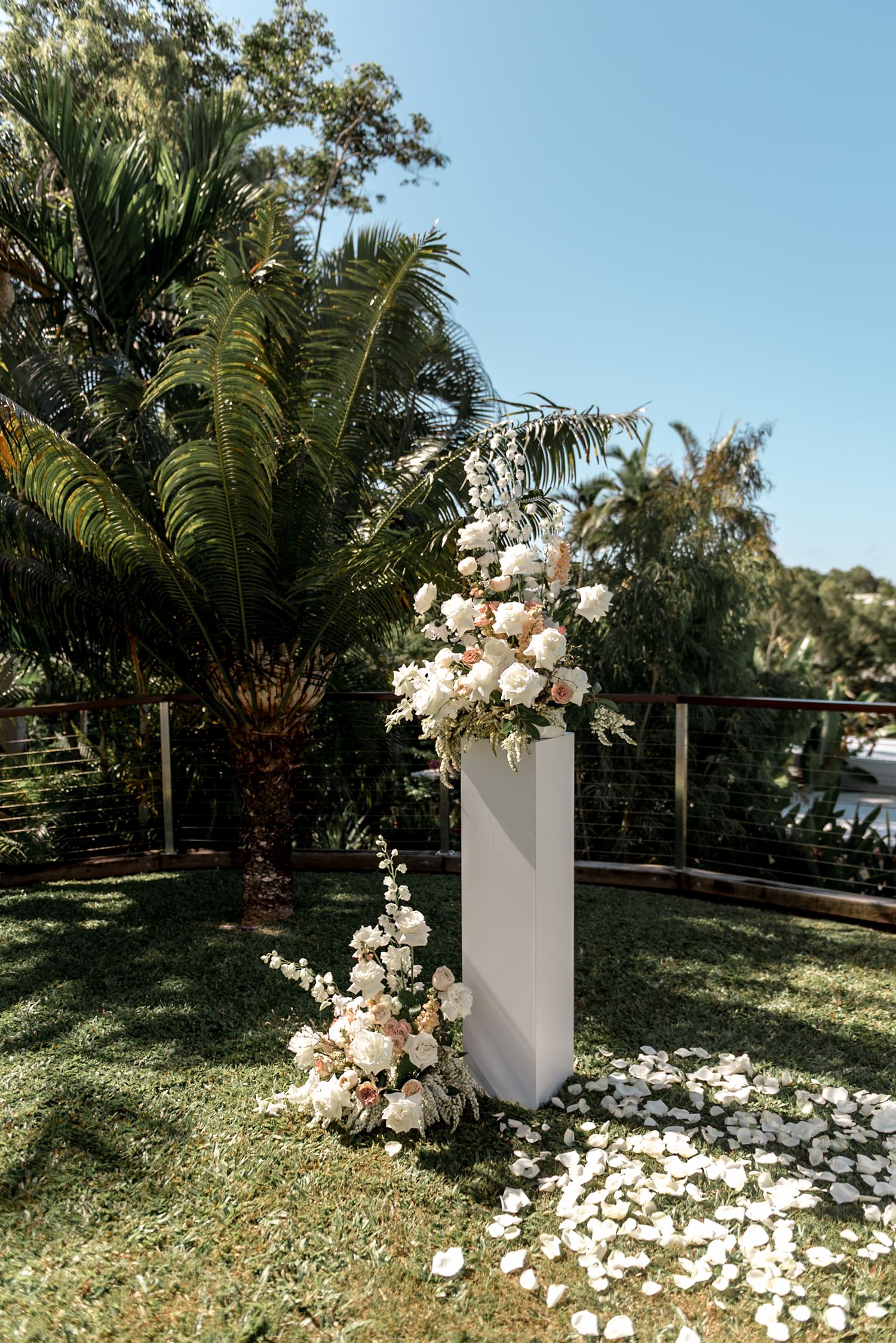 White wedding ceremony pillar adorned with lush white and blush floral arrangements on a green lawn with tropical palms.