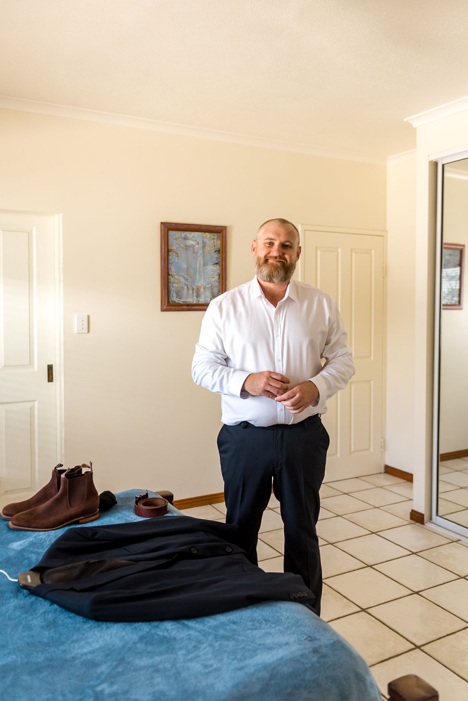 Bearded groom in white dress shirt preparing for wedding day in bright bedroom with suit on bed.