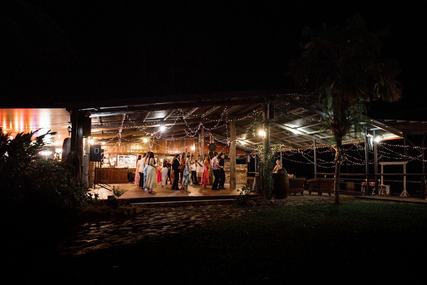 Rustic outdoor wedding reception venue illuminated with warm string lights at night, guests gathered under open-air pavilion.