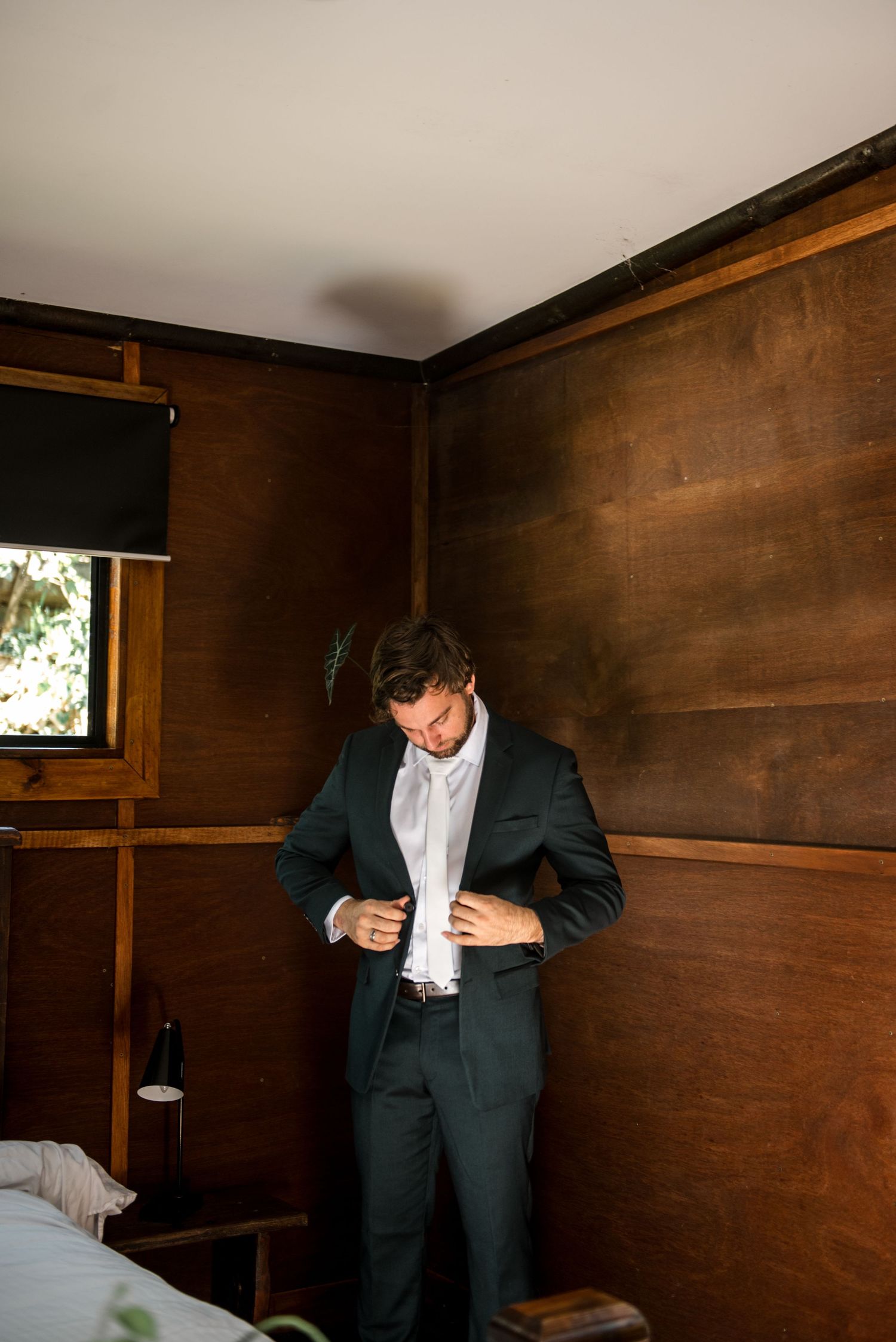 Groom in dark green suit buttoning his jacket in a warmly lit wooden paneled room.