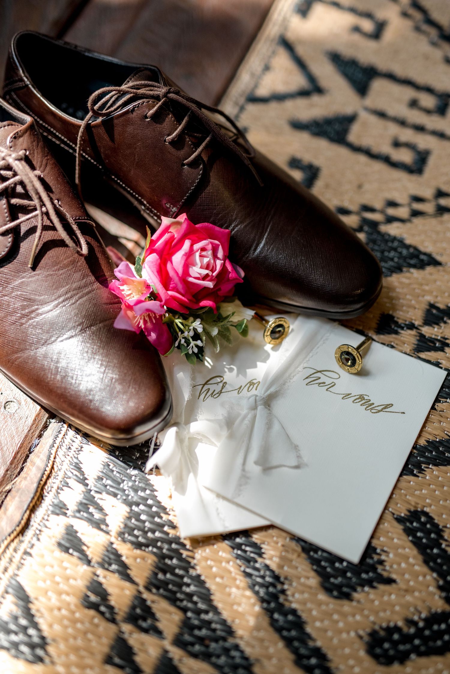Brown leather dress shoes, pink boutonniere, gold cufflinks, and wedding vow cards flat lay.